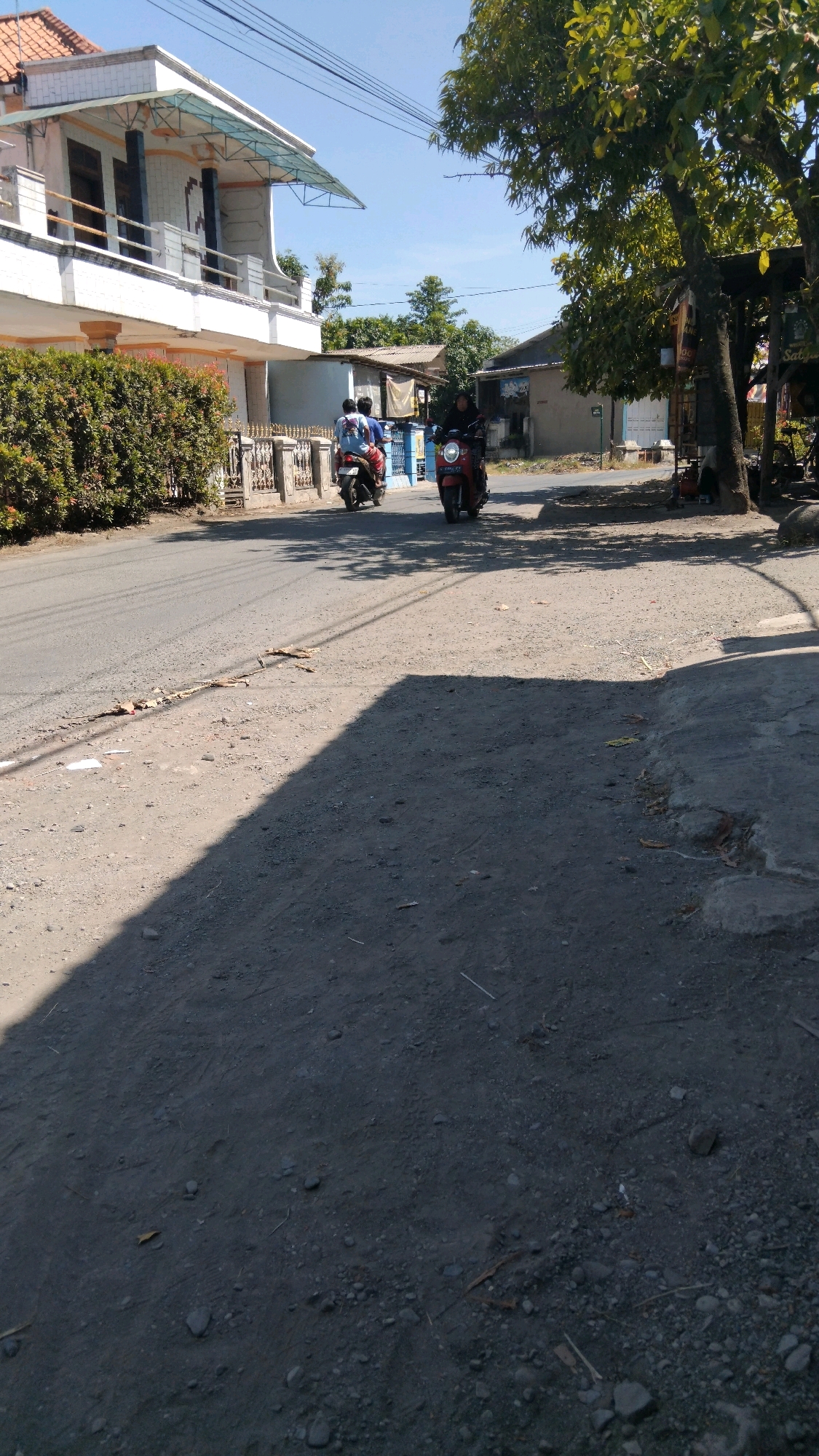 A street scene in Sidakaton, Indonesia, features a two-story building with white and orange accents and a blue-tiled roof on the left, partially obscured by a dense green hedge. Across the asphalt road, a tree with green foliage stands to the right, casting a shadow. Two individuals on a motorcycle are visible on the road. Further down the street, another person rides a red motorcycle. The ground is a mixture of asphalt and gravel. The sky is clear and blue.