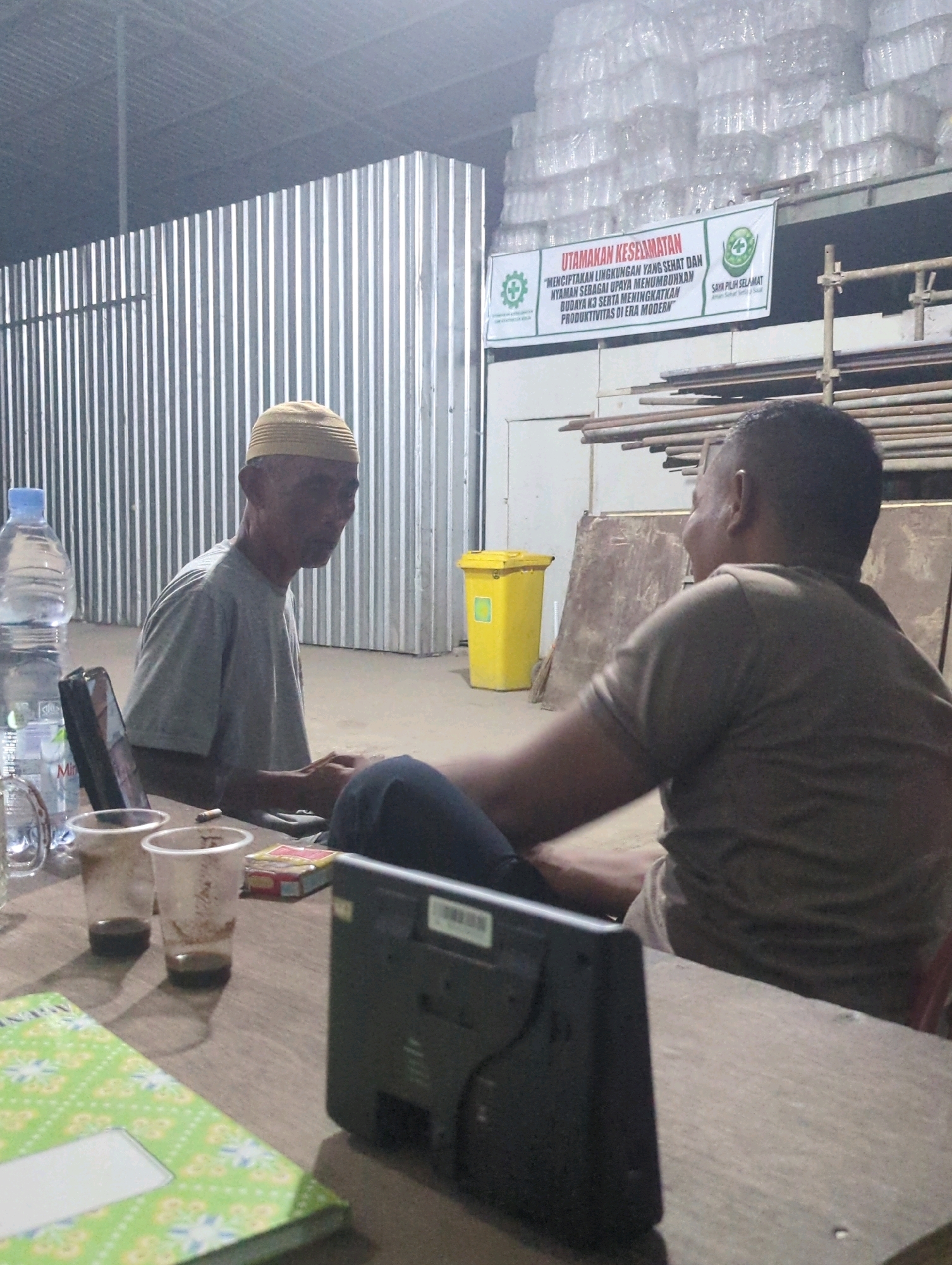 In a warehouse-like setting in Batu Belaman, Indonesia, two men are seated at a wooden table. The man on the left, wearing a yellow skullcap and a grey t-shirt, is looking towards the right. On the table in front of him are two plastic cups containing a dark liquid, a cigarette, and a pack of cigarettes. A bottle of water and a laptop are also visible. The man on the right, wearing a grey t-shirt, is facing away from the camera. A black electronic device is placed on the table in front of him. In the background, a yellow trash can and a corrugated metal wall are visible, as is a sign with text and logos. Stacks of transparent-wrapped items are visible on a shelf above.