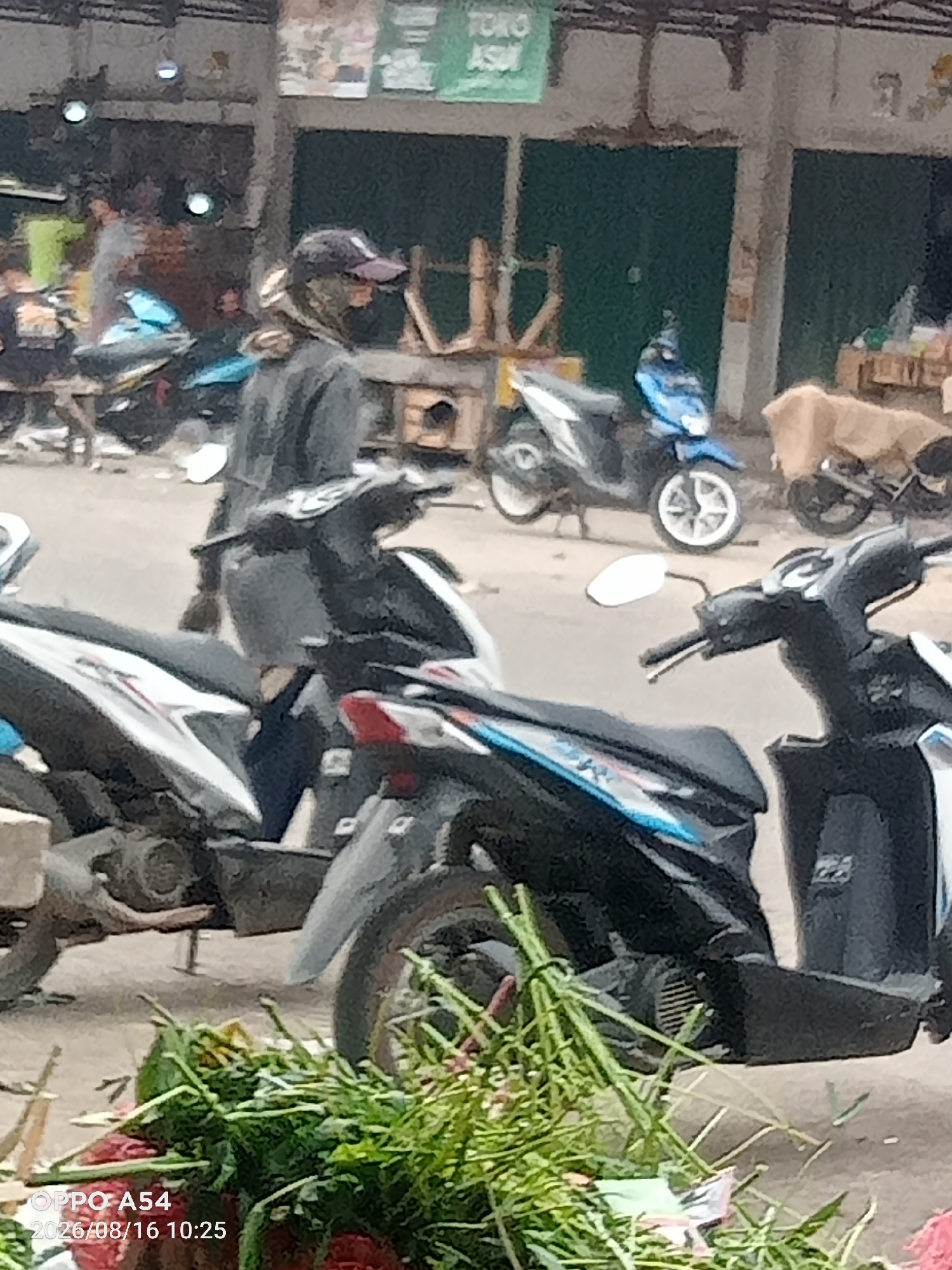 This is a daytime, eye-level, medium shot image taken outdoors in what appears to be a market setting. In the foreground, the rear portion of a black and silver motorcycle is visible, along with a pile of green foliage, possibly flowers or vegetables, to its left. Behind this, another black and white motorcycle is parked. Further in the background, a person wearing a dark hat and a gray jacket is standing next to a blue motorcycle. Several other motorcycles are parked in the vicinity. The background shows stalls with green metal doors and some wooden structures. The overall environment suggests a busy street or marketplace in Jambi, Indonesia, given the timestamp on the image: "2026/08/16 10:25".