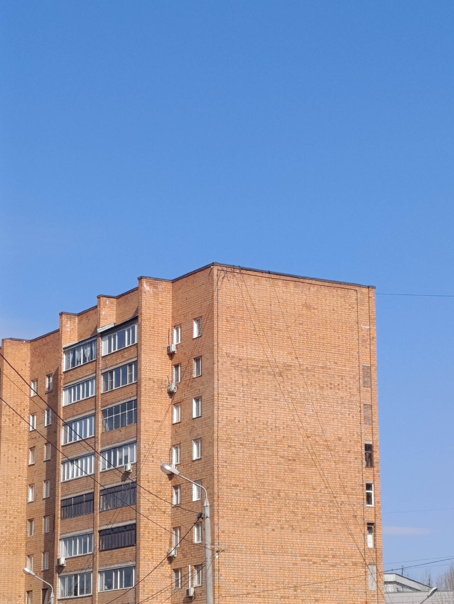 The image shows a multi-story brick apartment building against a clear blue sky. The building appears to be located in Krasnoyarsk, Russia. The building has multiple rows of windows and balconies. There are external air conditioning units attached to the building facade. Power lines are visible in the foreground, obscuring some of the building. A streetlight is visible near the building. The building's exterior is made of orange-brown bricks.