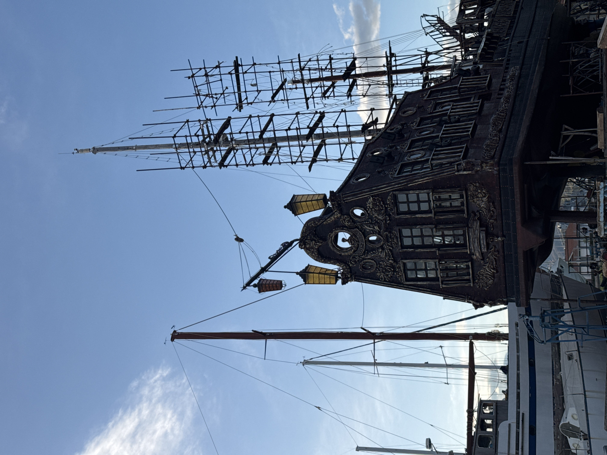 The image captures a partial view of a large, intricately decorated pirate ship. Its ornate design includes numerous windows, decorative carvings, and masts under construction. The sky is light blue with scattered clouds. The scene appears to be in a harbor, with other boats visible beside the pirate ship. The location is the city of Bar, Montenegro.