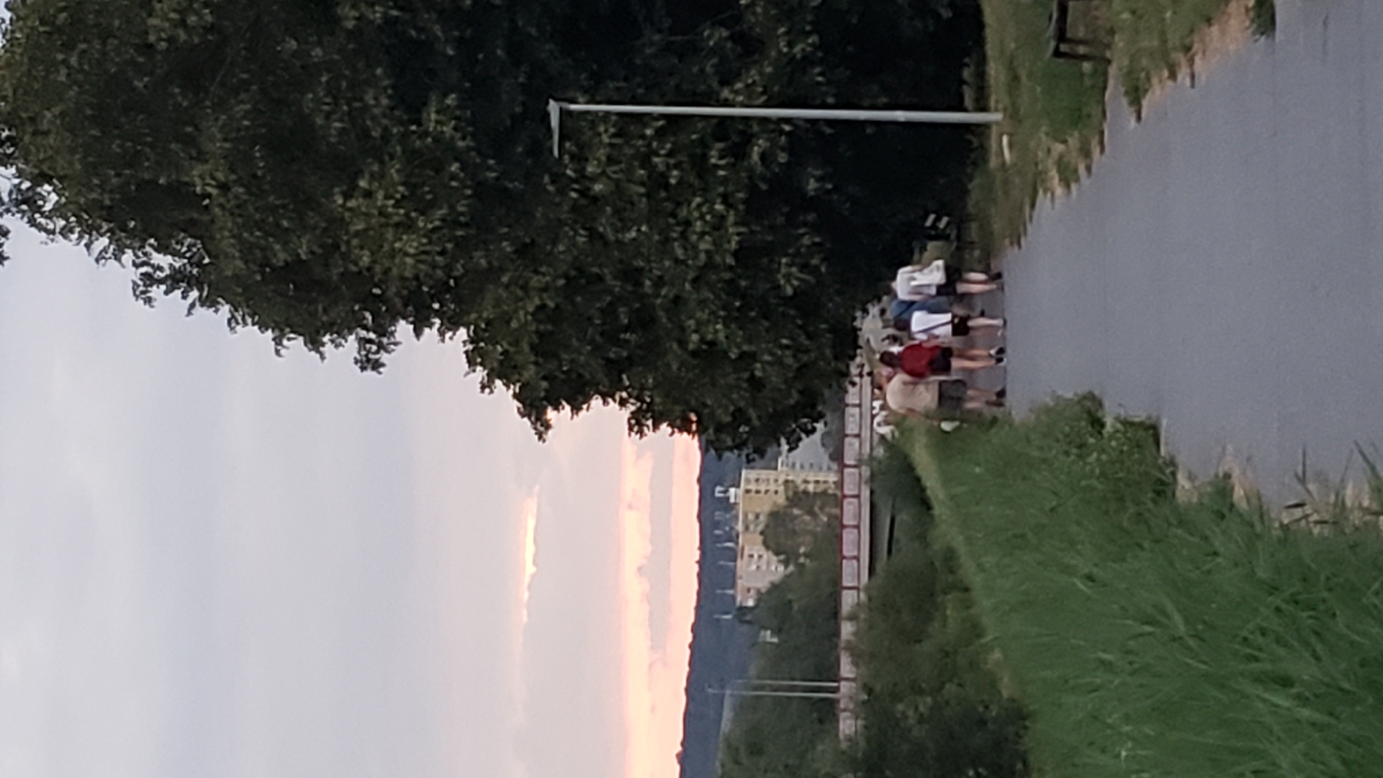 In Prešov, Slovakia, a group of five individuals walks away from the camera on a paved path adjacent to a grassy embankment. The sky exhibits soft pink and yellow hues, indicative of sunset or sunrise, with scattered clouds. A large, dark green tree dominates the right side of the frame, partially obscuring a modern street lamp. In the background, a distant cityscape features a multi-story building and a red-railed bridge spanning a body of water. The individuals are dressed in casual summer clothing, and their formation suggests a leisurely stroll.