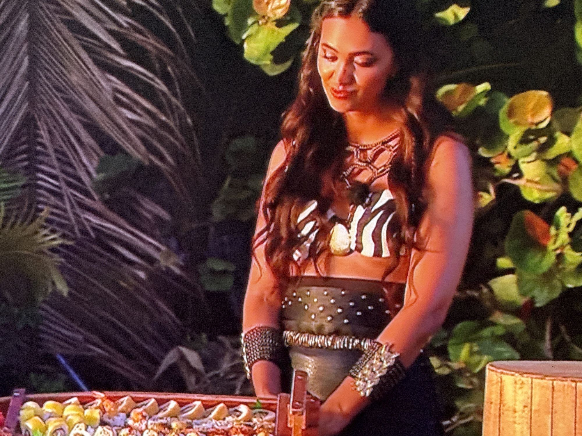 A medium shot captures a woman standing next to a table laden with sushi. She is dressed in what appears to be a tribal or themed costume, featuring a zebra-striped top, a dark, dotted waist belt, and armbands with elaborate designs. Her long, dark, wavy hair frames her face, which is turned slightly downwards, and she has a subtle, gentle expression.
The environment is lush and tropical, with broad green leaves of various plants forming a backdrop, along with a large palm frond to the left. The sushi on the table is presented in an orderly fashion, suggesting a catering setup. A round, bamboo-like structure is visible on the right side of the frame.
Location: Unknown, Unknown.