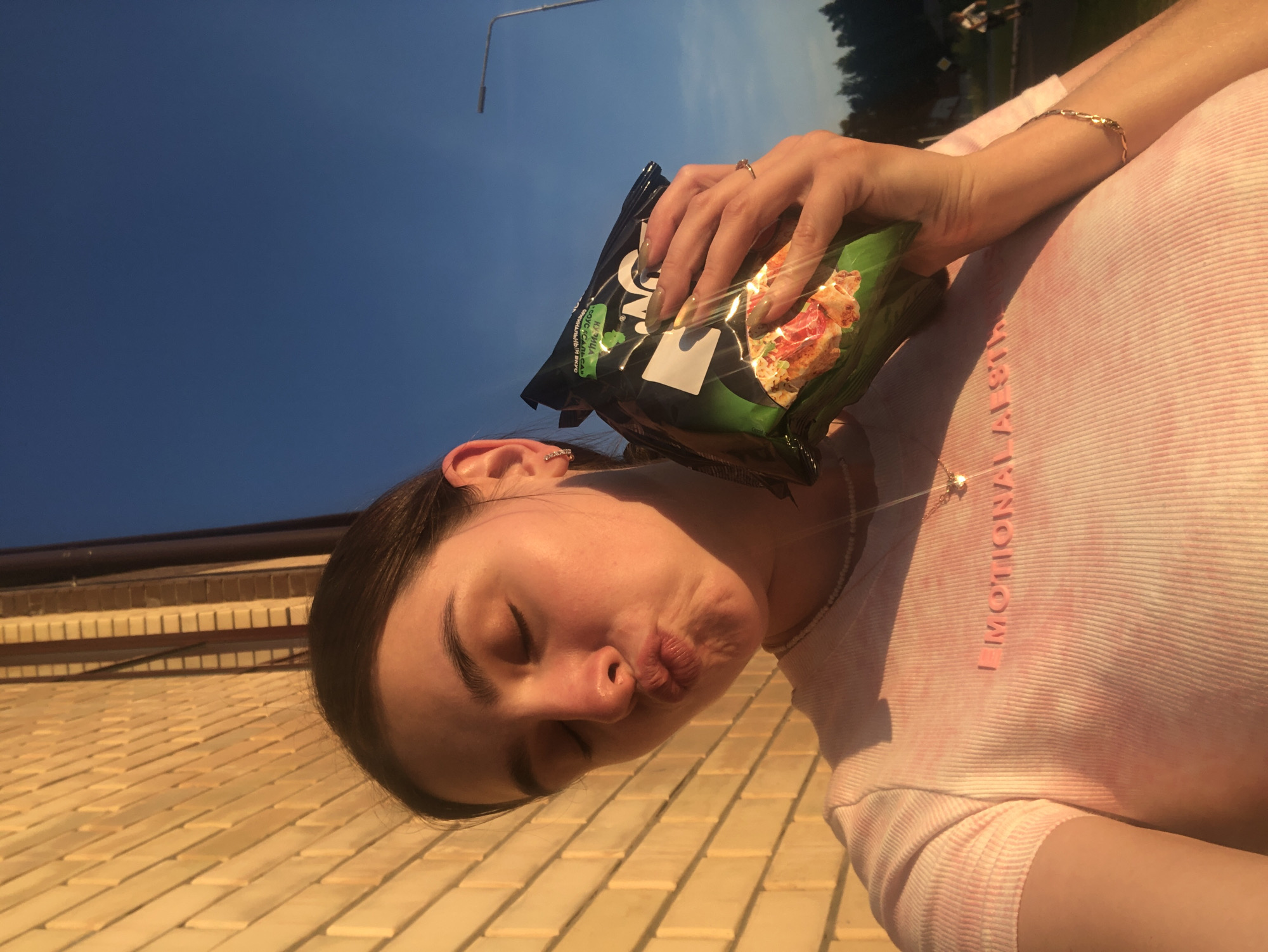 A young woman, with her eyes closed and lips puckered, holds a bag of chips in her right hand. She is wearing a pink tie-dye t-shirt with the word "EMOTIONAL" printed on it in pink lettering. A delicate necklace with a star pendant hangs around her neck. She has a ring on her right ring finger and a bracelet on her right wrist. The background consists of a brick wall on the left and a clear blue sky on the right. A street lamp and trees are visible in the distance. The scene appears to be outdoors, possibly in a residential area or park in Noviy, Russia, given the presence of a street lamp and the general setting. The lighting suggests it is late afternoon or early evening, with the sun casting a warm glow on the brick wall and the woman's face.