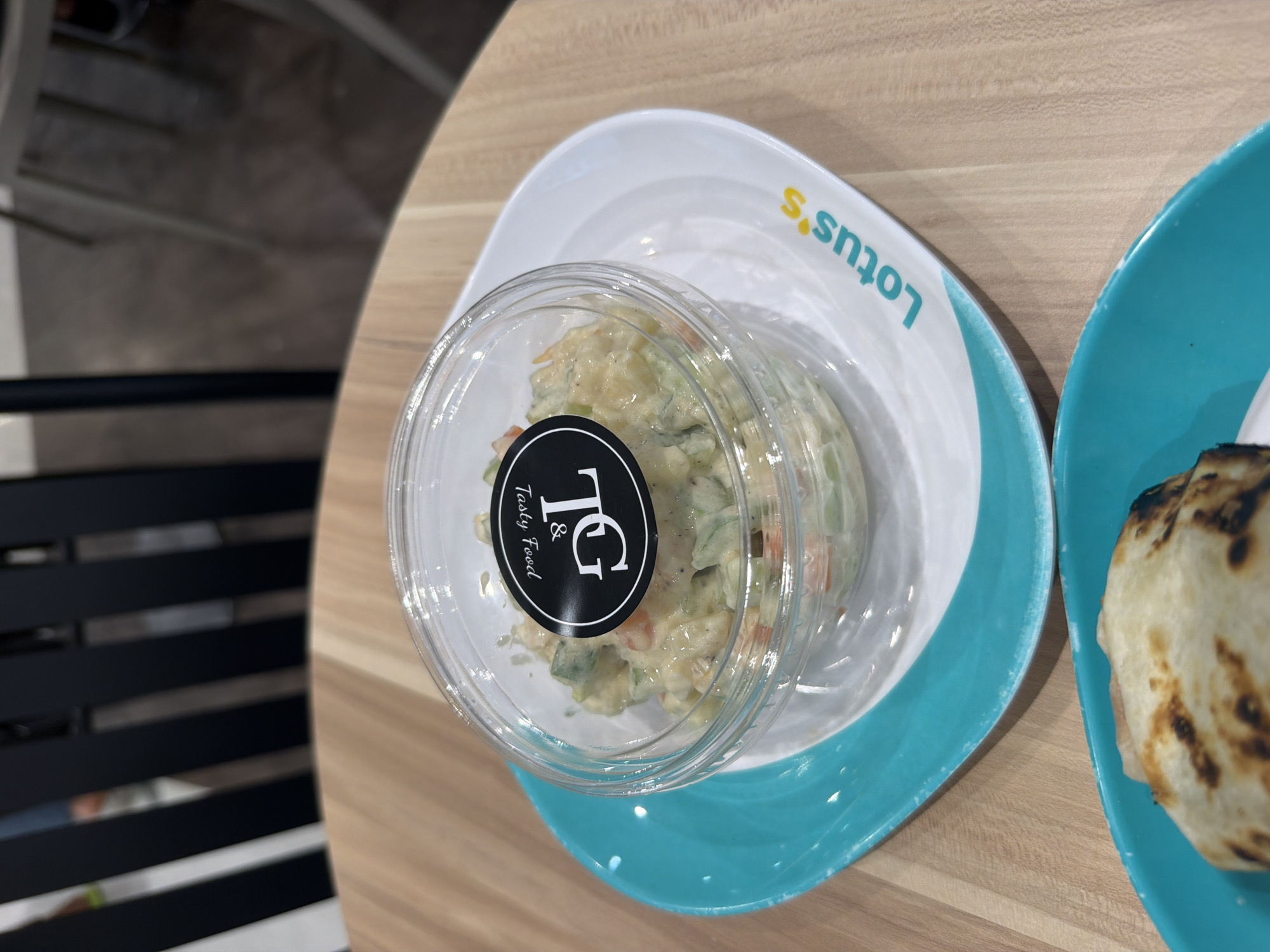 A close-up overhead shot shows a small, clear plastic container of coleslaw resting on a white plate with a teal rim. The plate, in turn, is placed on a light wood-grain table. A teal-rimmed plate with a portion of a grilled item is visible in the foreground, also on the table. A black chair is partially visible in the background. The coleslaw container has a black circular sticker with the text "& DL Tasty Food" and a stylized logo. The white plate also has the word "Lotus's" printed in teal and yellow. The image appears to be taken in a casual dining setting in the city of Ban Rang Rak, Thailand.