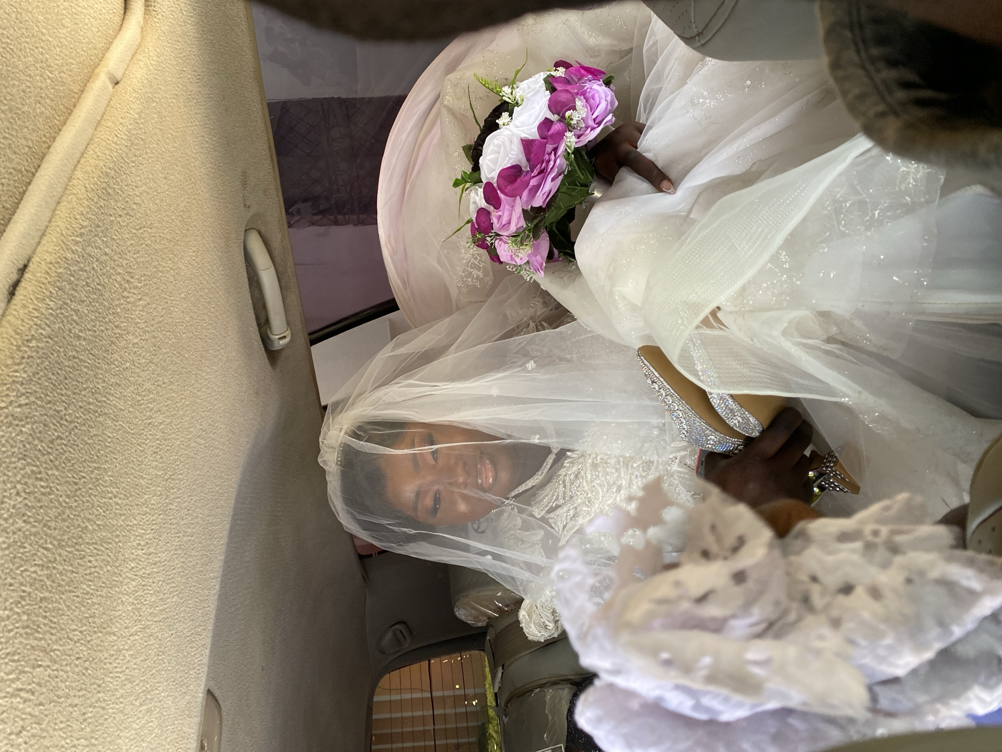 Inside a vehicle with a beige interior, a Black woman wearing a wedding dress and veil is holding a bouquet with white and purple flowers. The location appears to be Vom, Nigeria.