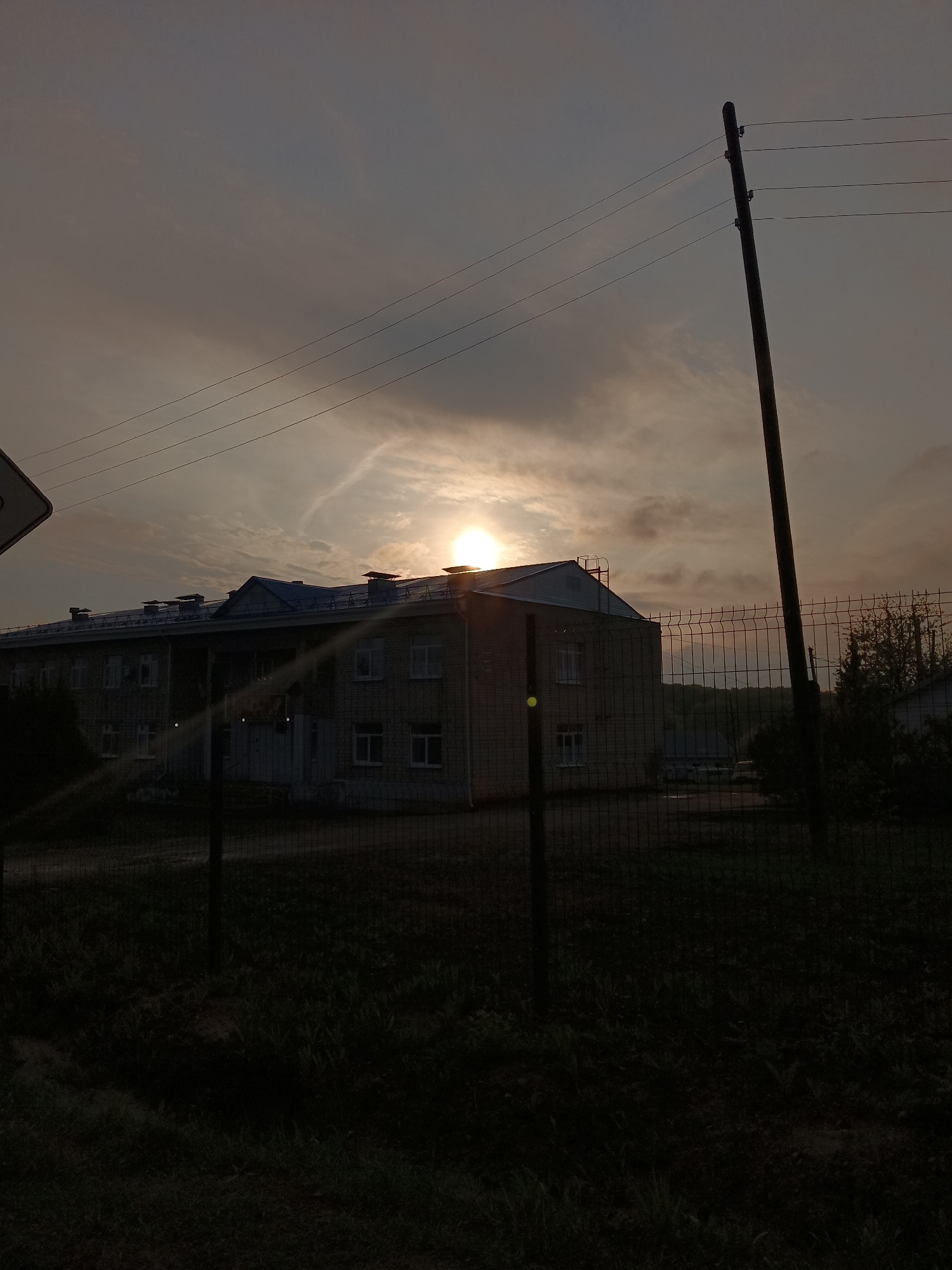 The image depicts a two-story brick building with a blue roof in the city of Фоки, Russia, partially obscured by a chain-link fence. The sun is setting behind the building, creating a silhouette effect. A wooden utility pole stands to the right of the building. The foreground is dark, with vegetation visible through the fence. The sky is cloudy.