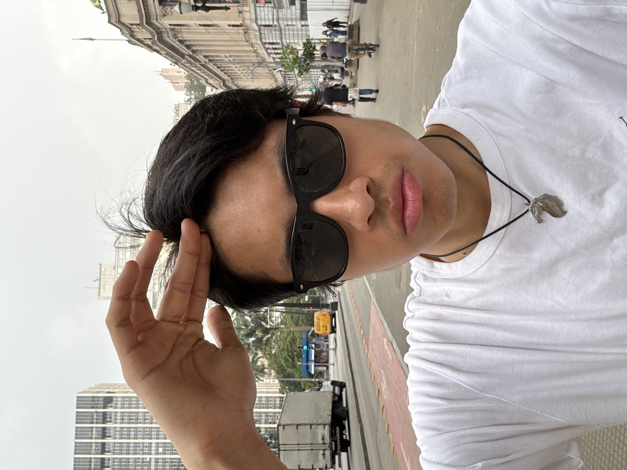 A selfie-style portrait captures a young person, likely male, in an urban setting identified as São Paulo, Brazil. The image is taken at a slight upward angle and is rotated 90 degrees counter-clockwise, showing the person on the left side of the frame looking towards the right. They have dark hair, dark sunglasses, and are wearing a plain white t-shirt. A silver pendant, resembling an animal's head (possibly a horse), hangs from a black cord around their neck. One hand is raised, casually touching their hair or the side of their head near the sunglasses. Their lips are slightly parted.
The background reveals a bustling city street under an overcast, grey sky. To the person's left and in the distance, a large, ornate historical building with columns and intricate details is visible, adorned with a Brazilian flag on a flagpole. Further back and to the right, modern high-rise buildings suggest a blend of old and new architecture. The street itself features traffic lights, streetlights, palm trees, and various vehicles including a white truck and what appears to be a yellow bus or taxi. Pedestrians can be seen in the distance, indicating a lively urban environment. The lighting is soft, consistent with a cloudy daytime setting. No discernible text is visible in the image.