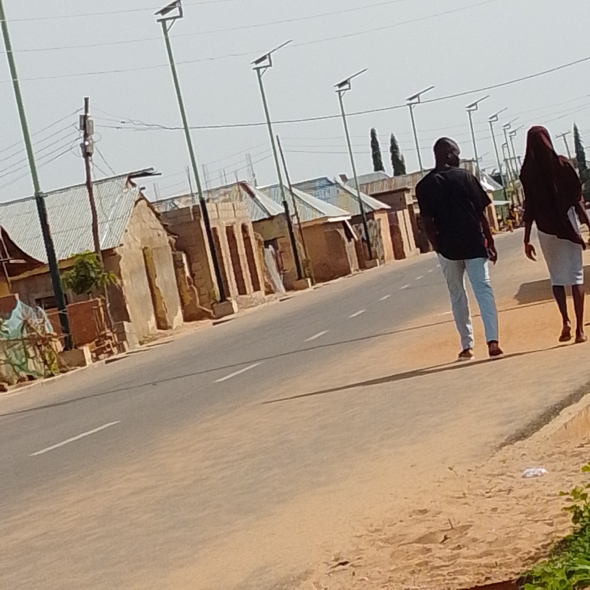 This media file captures a scene on a dusty road in Gombe, Nigeria, likely during the day under clear skies. Two figures are walking away from the camera along the right side of the road. The person in front is wearing a black t-shirt and light-colored pants, while the person behind them is covered in a dark, flowing garment, possibly a hijab. Their presence suggests an everyday activity, such as commuting or a leisurely stroll.
The road itself is paved with some white lane markings visible, indicating it's a well-traveled route. The left side of the road features a row of modest, single-story buildings with corrugated metal roofs, characteristic of many residential or commercial structures in the region. Several solar-powered streetlights line the side of the road, suggesting ongoing infrastructure development or a focus on renewable energy. A utility pole with wires is also visible, suggesting the presence of electricity. The overall impression is one of a typical day in a developing urban or semi-urban area. The sandy soil and the style of the buildings provide clear location cues to Gombe, Nigeria. There is no visible text in the image.