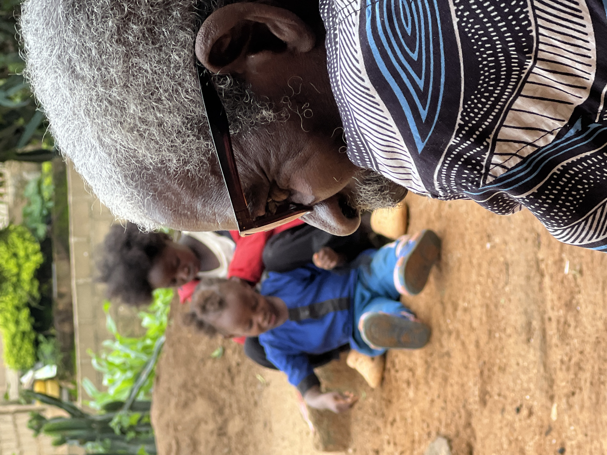 An elder with grey hair and glasses is in the foreground, facing away from the camera. Behind them, two children are sitting on the ground. One child, in a blue shirt and pants, is looking down at their hands. The other child, partially obscured, is wearing red and white. They are outdoors, with a sandy ground and some greenery in the background. The scene appears to be in a rural or semi-rural setting, possibly within a compound or yard. The image does not provide any specific landmarks or contextual clues to pinpoint the location within Jos, Nigeria.