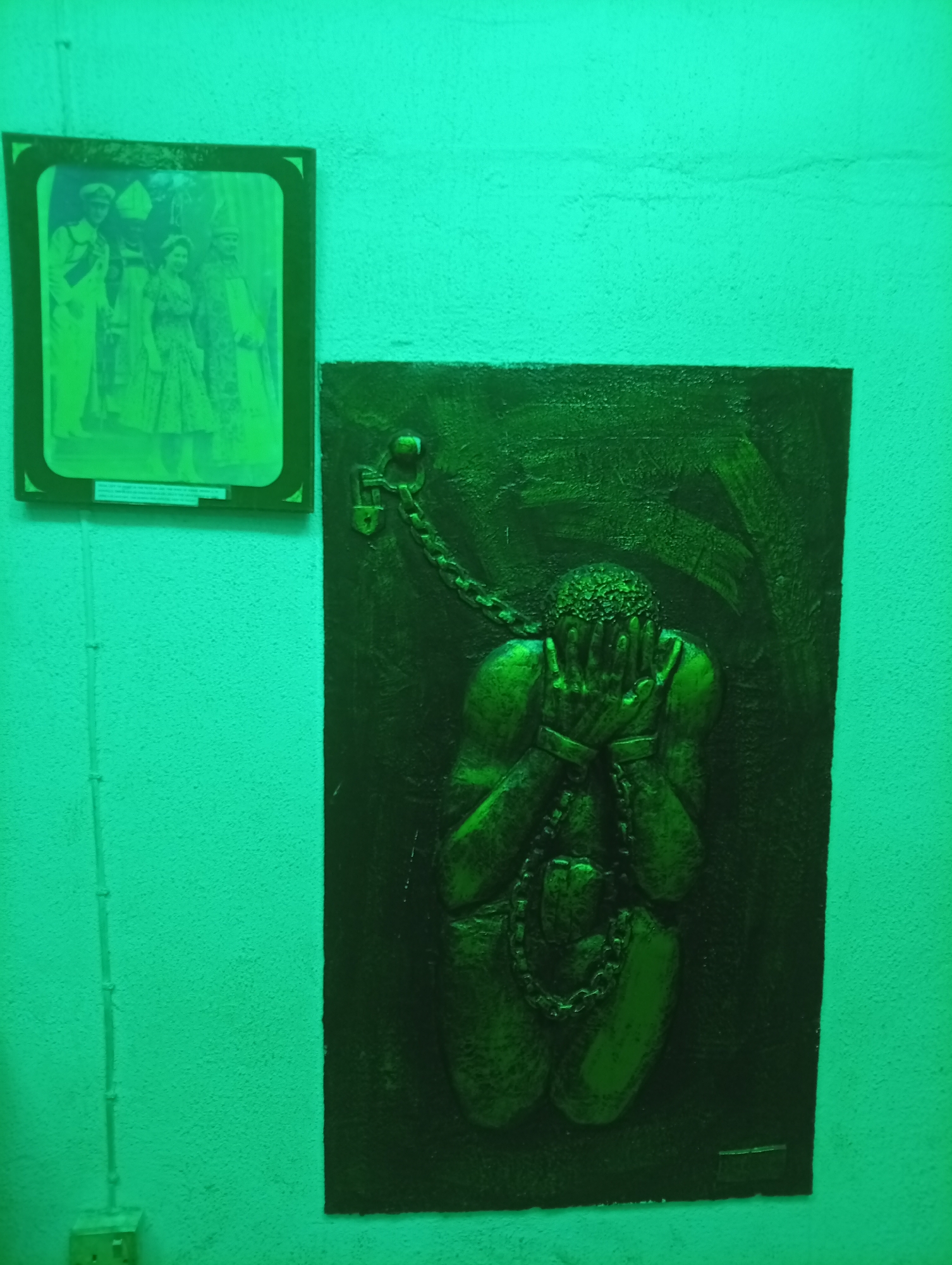 The image shows a section of a wall in what is likely a museum or historical site in Badagry, Nigeria. Two framed objects are visible: On the left is a framed photograph featuring multiple people, including one wearing what appears to be a papal mitre and another figure in a ceremonial robe, possibly of royalty. To the right is a relief sculpture depicting a human figure bound in chains. The figure has their head in their hands, suggesting despair or oppression. The chains extend from the figure's wrists and neck, with a padlock at the top of the sculpture. The lighting is predominantly green. A white electrical conduit runs along the wall to the left of the framed photograph.
