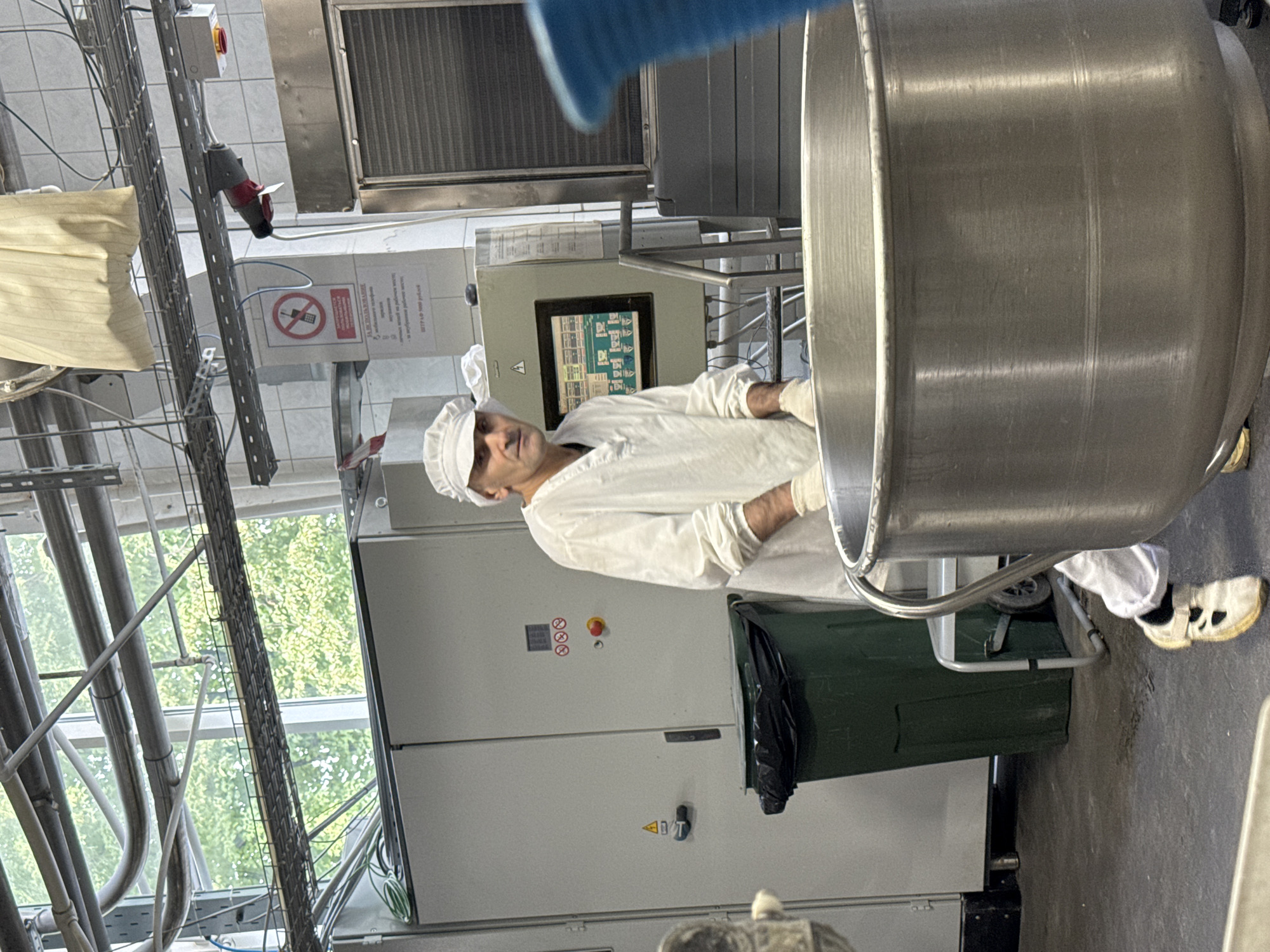 A person dressed in a white uniform and cap, wearing white gloves, stands in a food processing facility. The individual is positioned in front of a large, stainless-steel mixing bowl, which occupies the foreground of the image. To the left of the bowl, a dark green trash bin with a black liner is visible. Behind the trash bin are large, grey electrical cabinets. A monitor displaying technical information is mounted on a wall to the right of the cabinets. Industrial piping and shelving are visible overhead, with a window in the background revealing greenery. The overall environment suggests a commercial kitchen or food production area, likely within an industrial setting. The location is identified as Dedovsk, Russia.