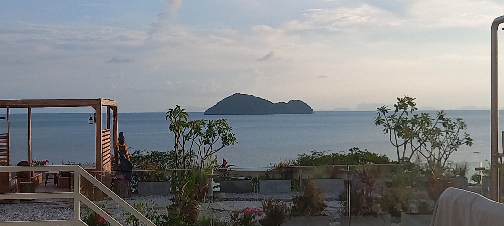 The image shows a view of the sea from what appears to be a raised patio or terrace in Baan Wok Tum, Thailand. In the distance, there is an island or landmass on the horizon, slightly obscured by haze. Closer to the viewer, a wooden structure resembling a pergola or gazebo with seating is visible. A statue stands near the structure. The foreground consists of a patio area with greenery, potted plants, and a glass railing. The sky is overcast with clouds. The general atmosphere of the scene is calm and serene.
