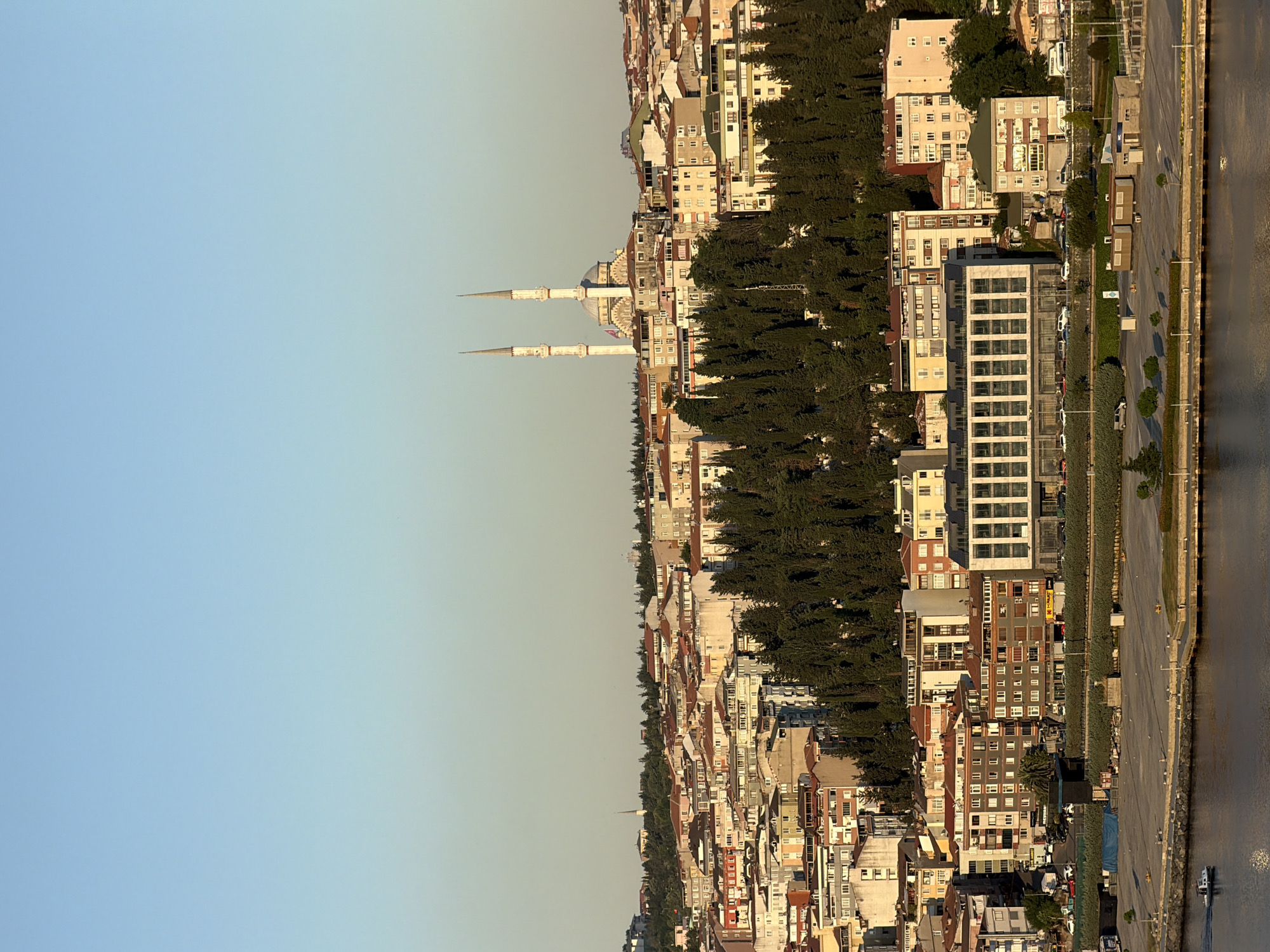 The image depicts a panoramic view of Eyüpsultan, Turkey, at golden hour. In the foreground, a calm body of water, likely the Golden Horn, reflects the warm light. A small boat navigates the water. Beyond the water, a large, paved area with minimal parked vehicles and scattered seabirds forms a waterfront promenade. Behind this, a dense line of cypress trees ascends a hill, creating a natural barrier between the waterfront and the urban landscape.
The hillside is densely populated with buildings of varying heights and architectural styles, characterized by red-tiled roofs and light-colored facades. Dominating the skyline of the urban area is a large mosque with a central dome and two prominent minarets, bathed in the warm evening light. Smaller residential buildings and commercial structures are interspersed throughout the cityscape, with some featuring balconies and multiple windows. The sky above is a clear, pale blue, transitioning to a lighter hue near the horizon, indicating the setting sun. The overall scene conveys a sense of a bustling urban environment integrated with natural elements and religious architecture, illuminated by the soft glow of dusk.
