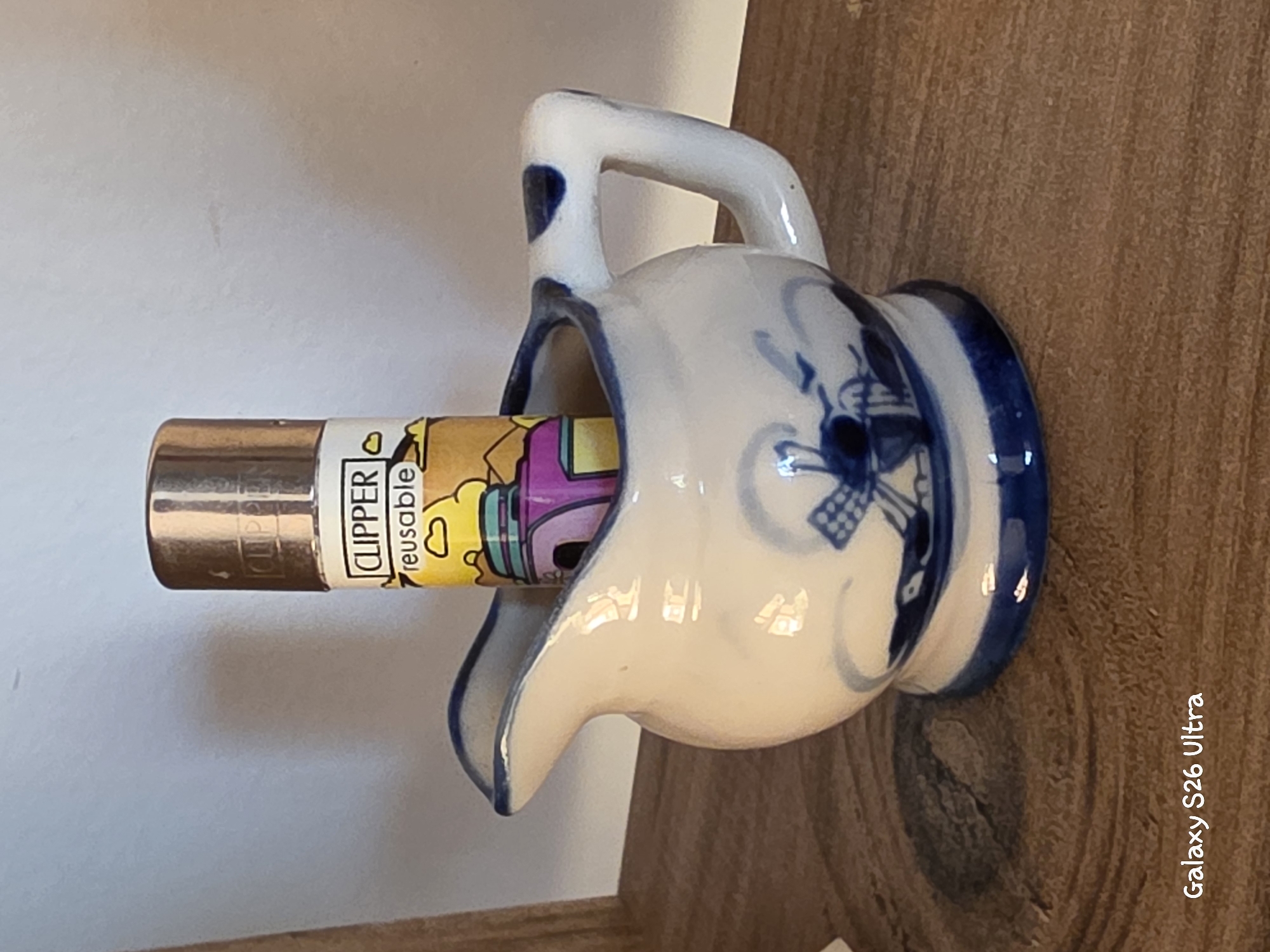 A decorative ceramic pitcher with blue windmill and floral motifs holds a Clipper brand lighter. The pitcher has a spout and a handle. The lighter has a metallic top and a colorful label that says "CLIPPER reusable." The objects are placed on a wooden surface. The image is taken at an angle, with the background appearing to be a white wall. The timestamp "Galaxy S26 Ultra" is visible in the bottom left corner. This scene is not located in the city of Plovdiv, Bulgaria.