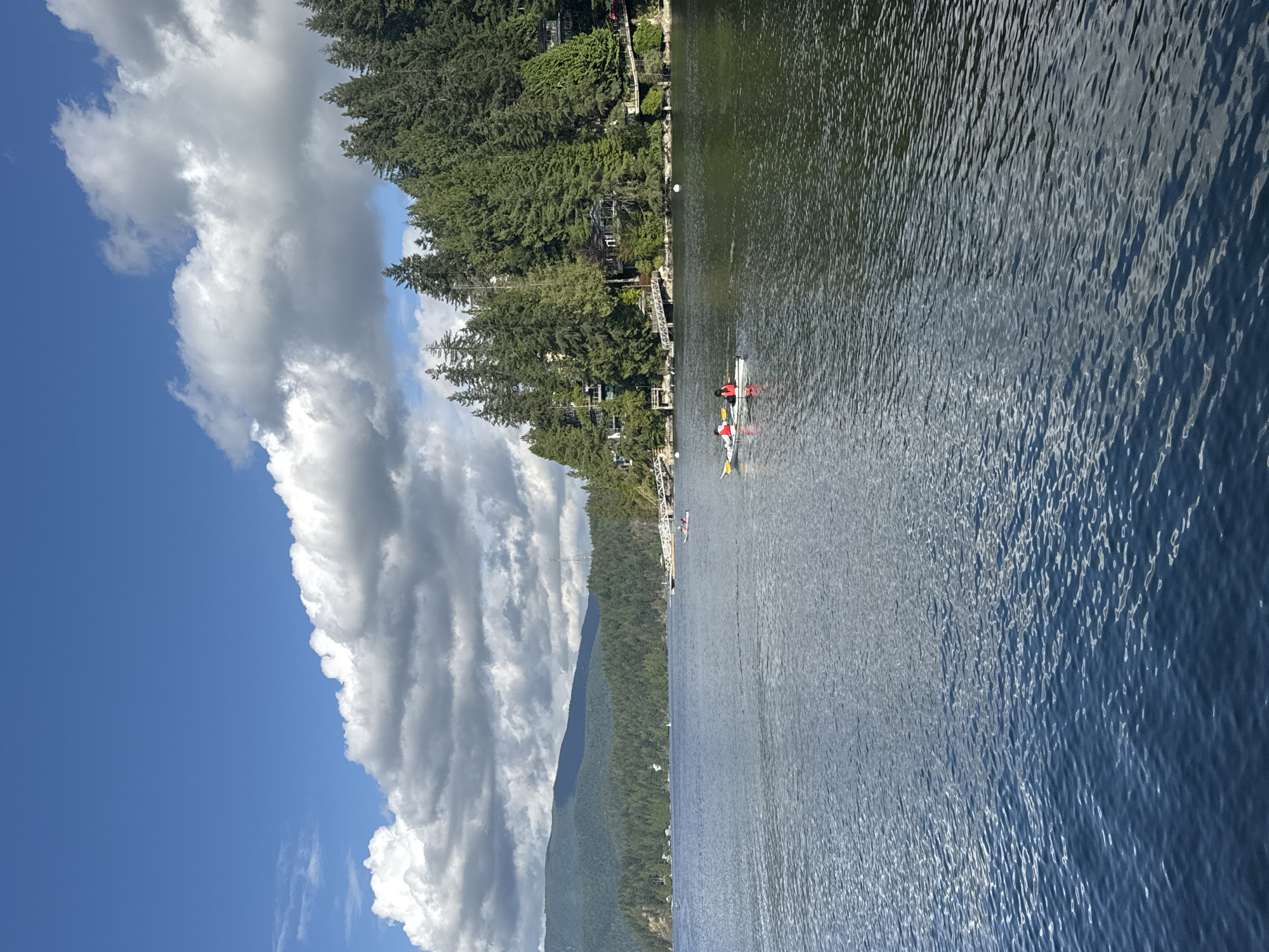 This image depicts a serene lake scene in the District of North Vancouver, Canada, under a partly cloudy sky. In the foreground, the deep blue water is textured with gentle ripples, reflecting the light. A two-person kayak is positioned in the middle ground, with two individuals, one in a red top and the other in a white top, paddling. Further back, a forested hillside rises, densely packed with tall evergreen trees. Several houses are nestled among the trees along the shore, some featuring elevated walkways and docks. A long pier extends into the water from the right side of the frame. In the distance, a range of mist-shrouded mountains forms the backdrop. The overall impression is one of natural beauty and peaceful recreation.