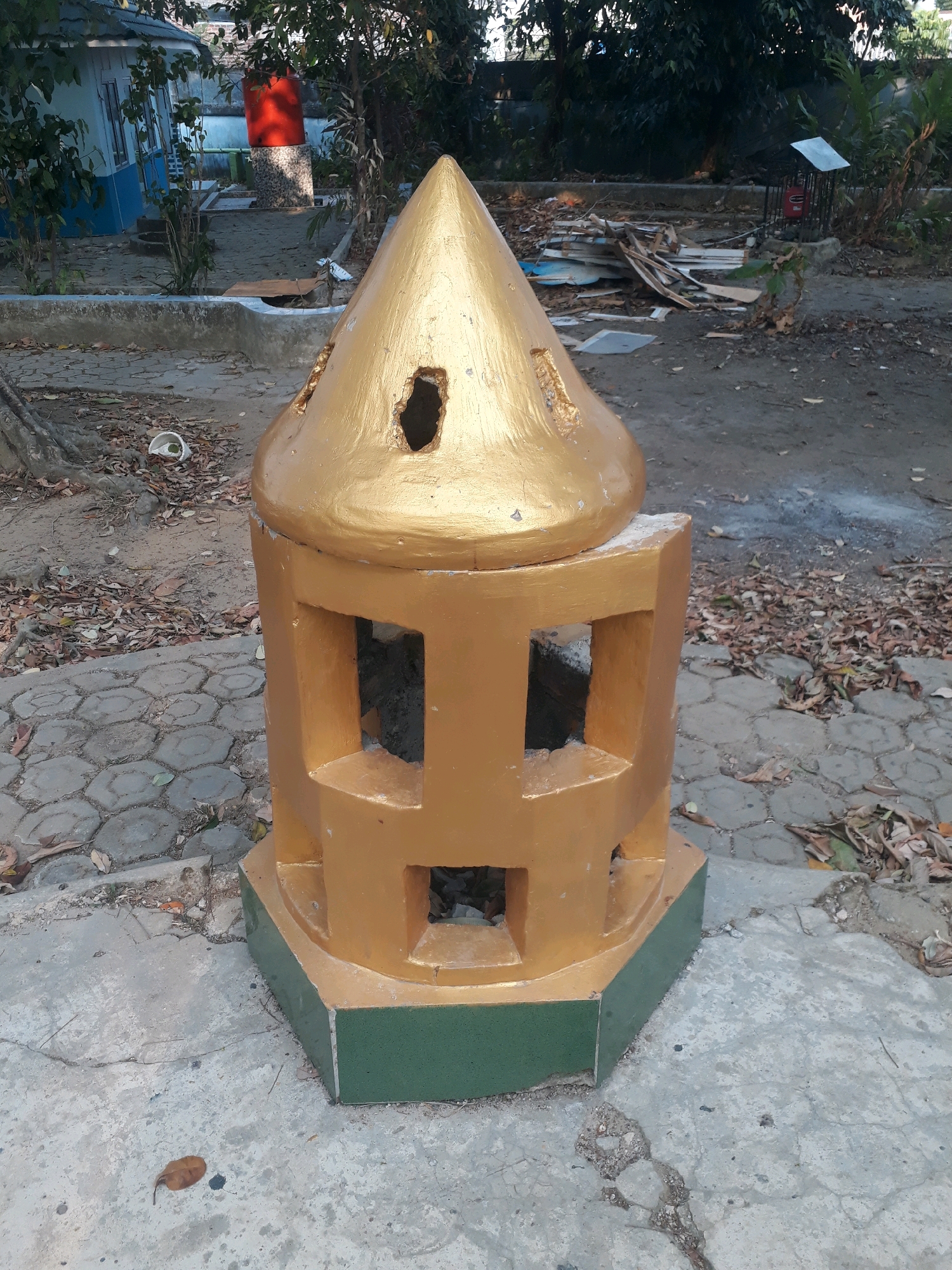 The image captures an outdoor scene, likely in the city of Pangkalpinang, Indonesia. In the foreground, a golden, conical structure with square openings stands on a concrete base painted green. The structure appears weathered and has some damage. Surrounding the structure is a paved area with hexagonal gray stones, interspersed with concrete. Fallen leaves are scattered across the ground, suggesting a natural or somewhat neglected environment.
In the background, a light blue building with white trim is visible, along with a large red water tank. The area also contains trees and shrubs. Further back, a pile of discarded wood and debris is present. The overall impression is of an outdoor space that may be part of a park, a public area, or a residential compound, possibly in a tropical climate given the vegetation. No people are visible in the image.