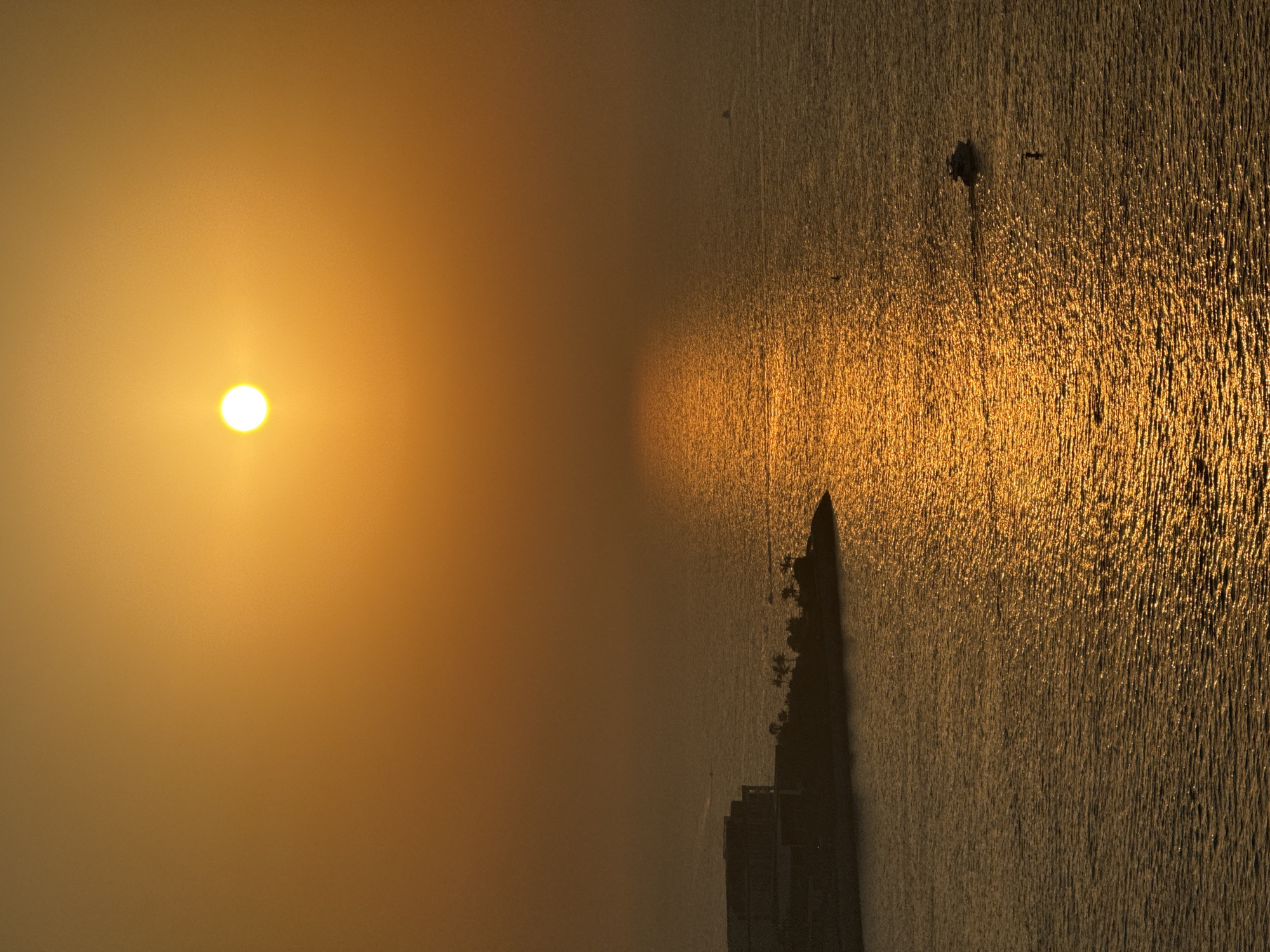This is an aerial vertical shot of the Persian Gulf in Dubai, UAE, during sunset. The sun, a bright, white orb, is positioned in the upper center of the frame, casting a golden, hazy glow across the scene. The water reflects the sun's light, creating a shimmering path that stretches from the horizon towards the viewer.
On the left side of the image, a man-made peninsula juts into the water. It is topped with several buildings, appearing as dark, angular structures against the warm sky. A scattering of palm trees can be seen on the peninsula. In the water near the peninsula, there are several small boats, one of which is moving through the water, leaving a visible wake. Another, smaller boat is further out in the water, and a single person can be seen on a paddleboard in the foreground. The overall atmosphere is serene, with the warm colors of sunset dominating the image.