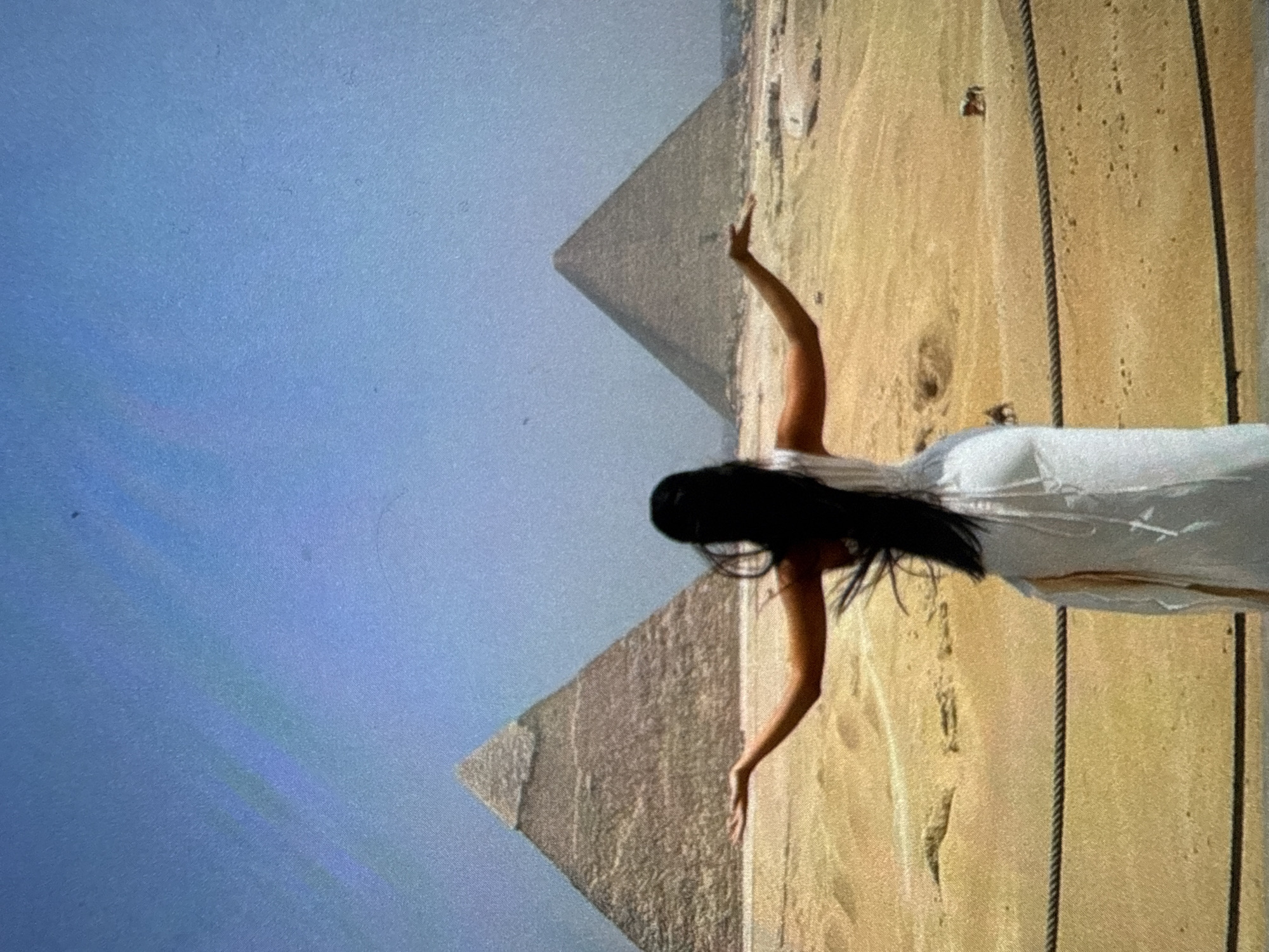 A photo depicts a woman in a white dress standing with raised arms against the backdrop of the Egyptian pyramids. The location is in Egypt, Giza, by the Great Pyramid of Giza.