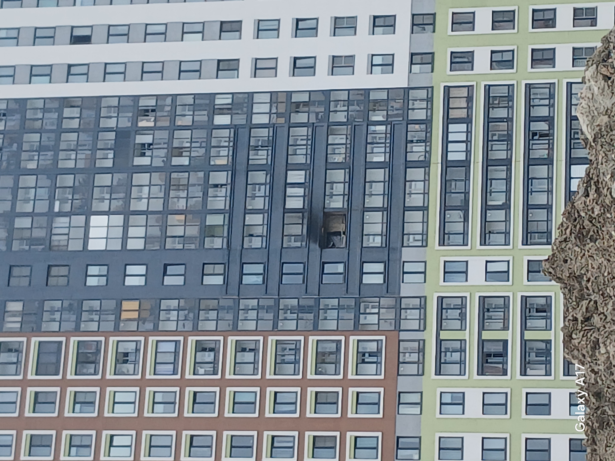 The image shows a multi-story building with a variety of window arrangements and facade colors in what is likely Yekaterinburg, Russia, given its architectural style. A single window in the central section of the building has smoke or soot damage, indicating possible fire damage. The building has several sections of different colors: white, grey, green, and brown. A blurred object, possibly a tree trunk, is present on the right side of the frame. The text "Galaxy A17" is visible on the bottom right, indicating the camera model used to take the picture. The building's exterior shows a repeating pattern of windows, suggesting an apartment or office structure.
