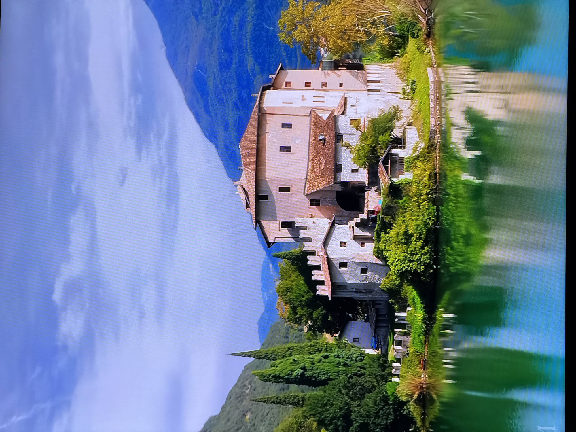 The image depicts a picturesque castle situated on a small island in a serene lake, surrounded by lush greenery and majestic mountains under a partly cloudy sky. The castle itself is a multi-story stone structure with a terracotta tiled roof, featuring multiple windows and battlements. The water of the lake is a vibrant green, reflecting the castle and the surrounding vegetation. The landscape includes tall, slender cypress trees and a large deciduous tree with yellowing foliage, suggesting an autumn setting. The mountains in the background are a deep blue, indicative of distance. The scene conveys a sense of tranquility and historical grandeur. This image is not located in Chirchik, Uzbekistan.