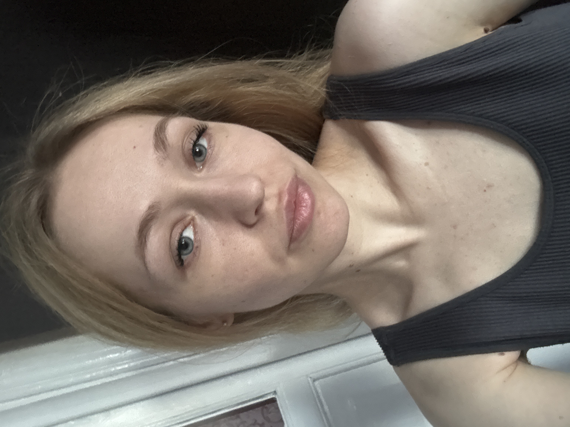 A young, fair-skinned woman with blonde hair and blue eyes is captured in a close-up, selfie-style shot. She is wearing a dark gray ribbed tank top. The background is softly blurred, with the left side of the image featuring a light-colored, paneled door and the right side a dark, indistinct area. The woman's facial expression is neutral. The lighting suggests an indoor setting, possibly a home.