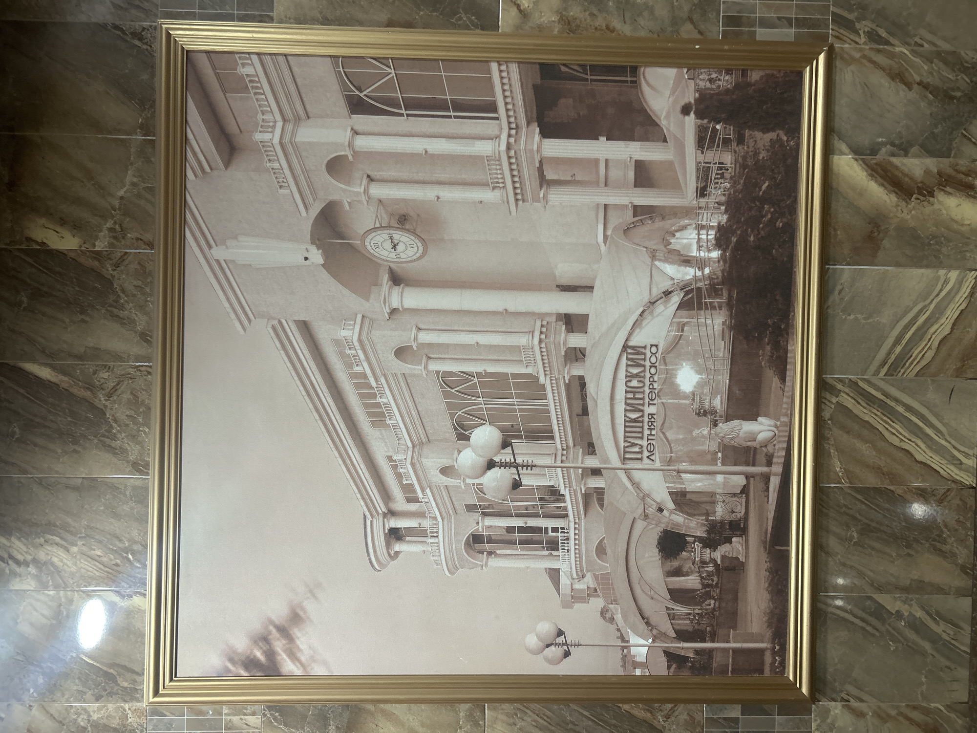 The sepia-toned photograph, framed in gold, depicts a grand building with classical architectural elements, including columns, arched windows, and balconies. A large clock is visible on the facade. In the foreground, there are illuminated street lamps and awnings over an outdoor seating area, suggesting a restaurant or cafe. The text visible on one of the awnings reads "ПУШКИНСКИЙ ЛЕТНЯЯ ТЕРРАСА", which translates to "Pushkinsky Summer Terrace". This indicates the location is the Pushkinsky Summer Terrace in Kursk, Russia. Several figures are visible, including what appear to be statues of lions and people walking. The building is set against a cloudy sky. The photograph is mounted on a wall covered with marbled tiles.