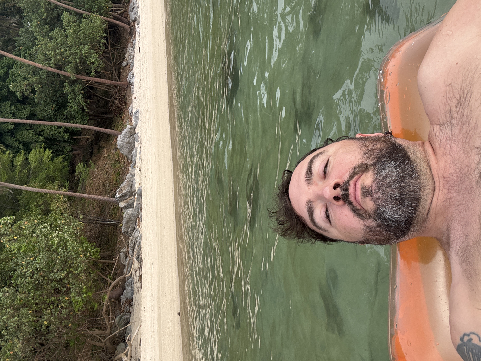 This media file captures a tranquil scene of a man relaxing in clear, calm water at a tropical beach in Baan Pang Ka, Thailand. The image is taken from a low angle, looking up at the man who is floating on his back, partially submerged, with his head and upper torso visible. He appears to be resting peacefully with his eyes closed, supported by an orange inflatable. He has dark, curly hair and a dark beard with noticeable grey streaks. His chest and arm are visible, showing body hair and a tattoo on his left shoulder/upper arm area.
The setting is a picturesque beach. To the left of the man, a narrow strip of light-colored sand forms the shoreline, bordered by various rocks. Beyond the sand, a dense line of lush green tropical vegetation extends, featuring several tall palm trees that lean gracefully towards the water. The water itself is a clear, greenish-blue, reflecting the light and showing gentle ripples on its surface. The bright, even lighting suggests it is daytime, likely a sunny and pleasant day. There is no visible text in the image. The overall impression is one of relaxation and serenity in a beautiful natural environment.
