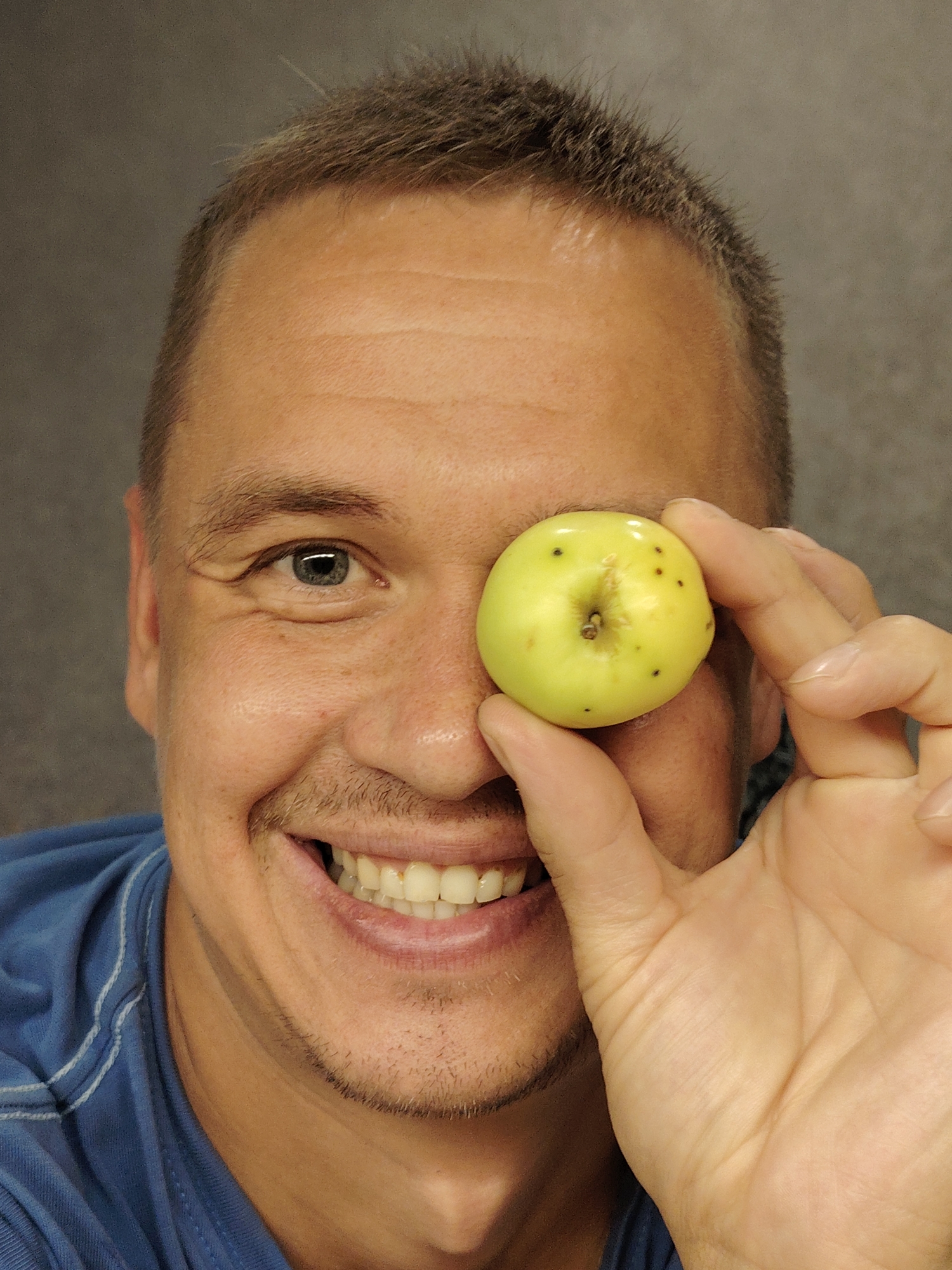 A man with short, dark-brown hair and light skin is holding a small, light-green apple in front of his right eye. He is wearing a blue shirt. He has a mustache and a light beard. The background is a plain, dark gray wall. The image was taken indoors.