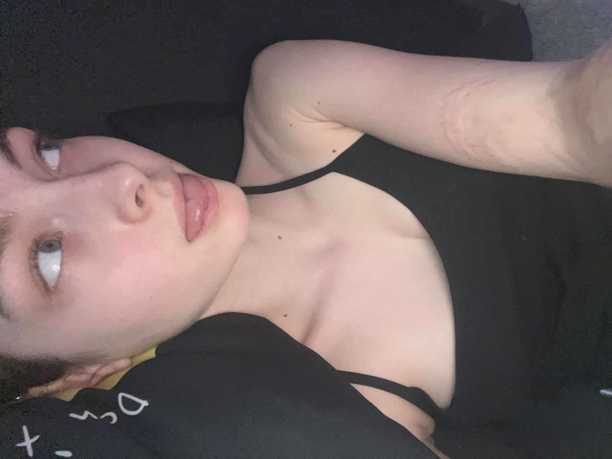 A fair-skinned person is lying down, their head tilted to the left. They are wearing a black top with thin straps. The person's tongue is slightly protruding. There are visible marks on their right arm. The background is dark. The location is not identifiable as Saint Petersburg, Russia, or any other specific location.