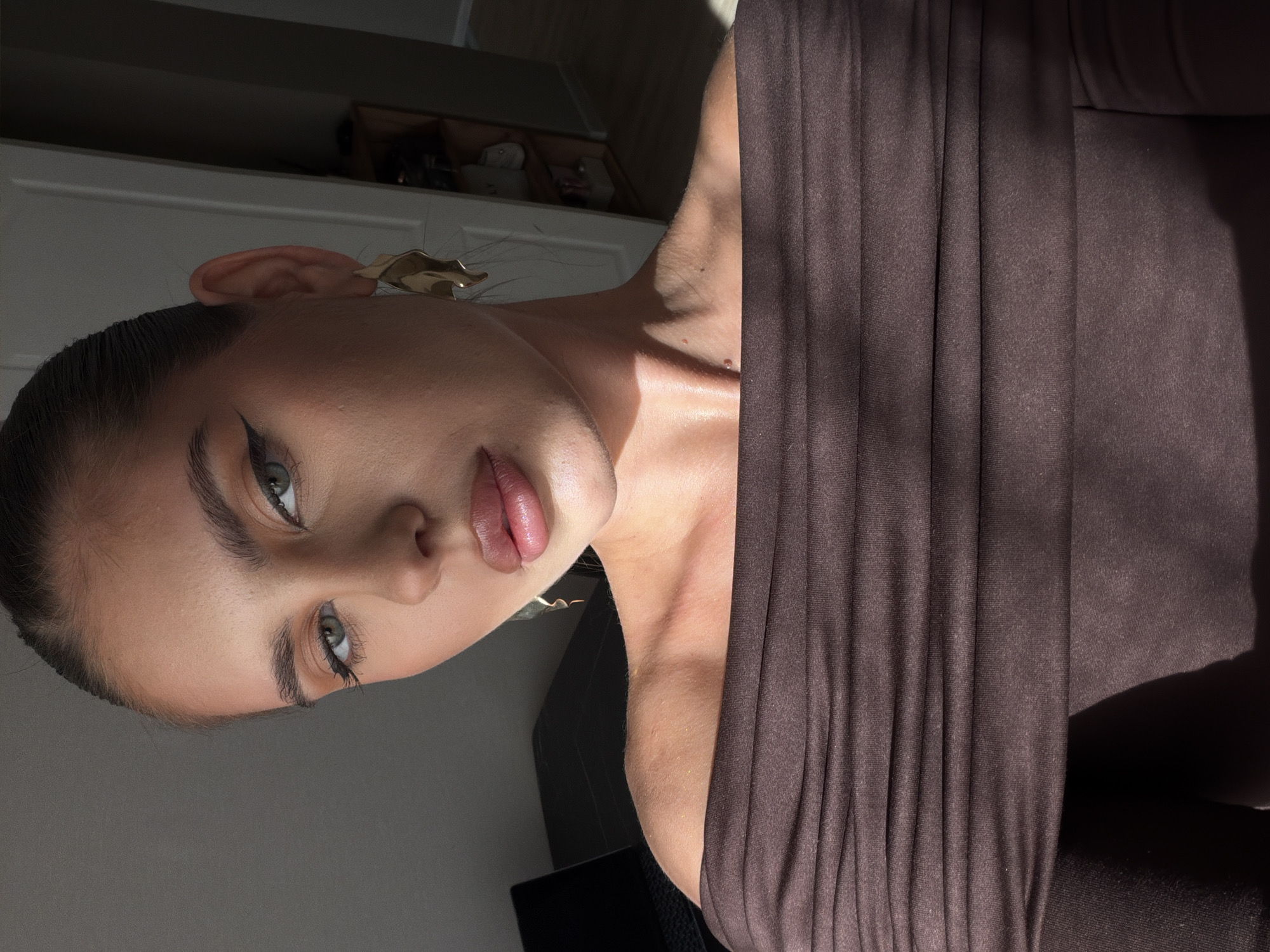 A portrait of a fair-skinned woman, likely in her late teens or early twenties, is oriented at an angle. She is wearing a dark brown, draped garment that occupies a significant portion of the right side of the frame. Her hair is pulled back, and she sports eyeliner. She is wearing earrings. The background features a white door and some items on a shelf. The photograph appears to be taken indoors with natural light. There is no clear indication of the location being Krasnoyarsk, Russia.
