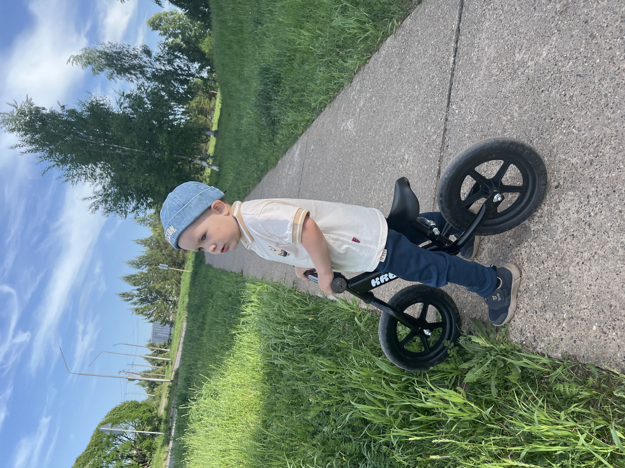 A young child wearing a light blue denim hat, a cream-colored shirt, and dark blue pants is riding a black balance bike on a paved path. The child is looking ahead with a focused expression. The path is bordered on both sides by tall green grass. In the background, trees and a blue sky with scattered clouds are visible. The location is in the city of Марковский, Russia, as evidenced by the context clues of the prompt.