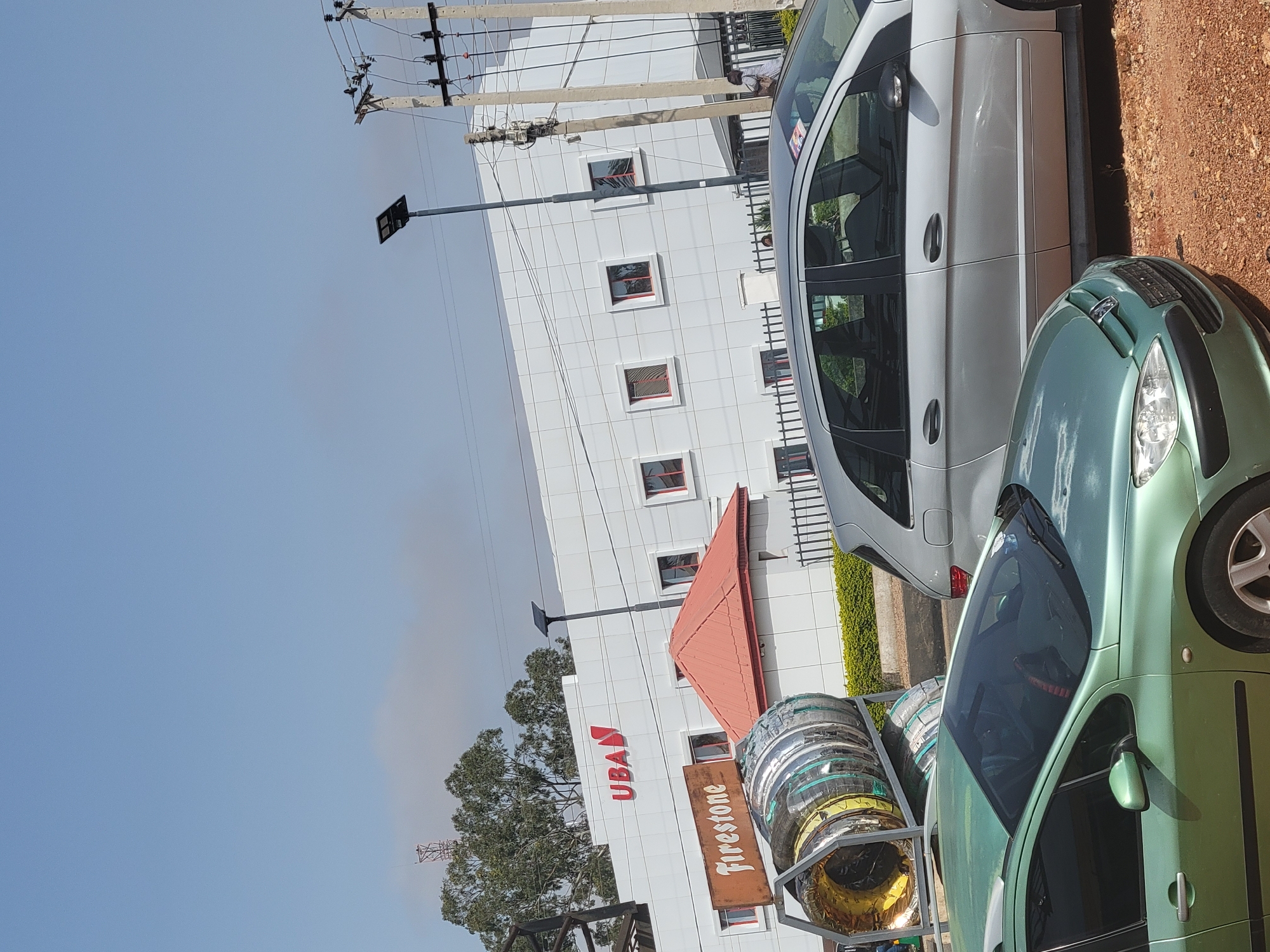 The image shows a street scene in an urban location. In the foreground, there are two cars: a light green car and a silver van. To the left, there is a stack of tires. Behind the vehicles is a white building with a red roof, and the "UBA" logo. A "Firestone" sign is visible next to the tires. In the background, there are trees and utility poles. The sky is light blue.