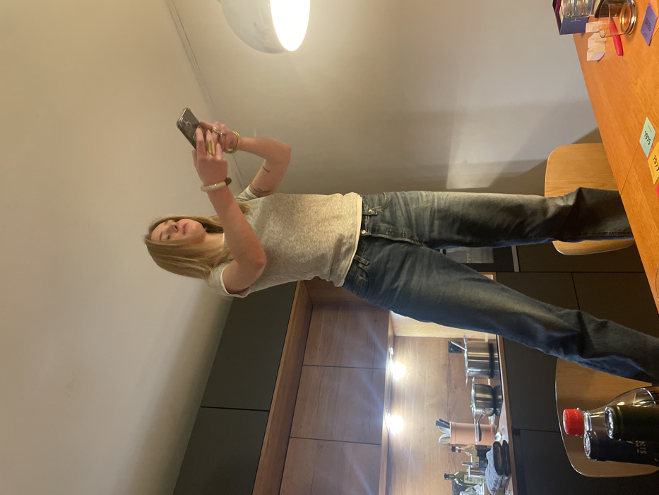 A woman stands in a kitchen, holding a smartphone. She has blonde hair and wears a gray t-shirt and jeans. Cabinets are visible behind her, as well as cooking utensils on the counter. A table with various items, including cards and a glass, is on the right side of the image. A light fixture is on the ceiling.