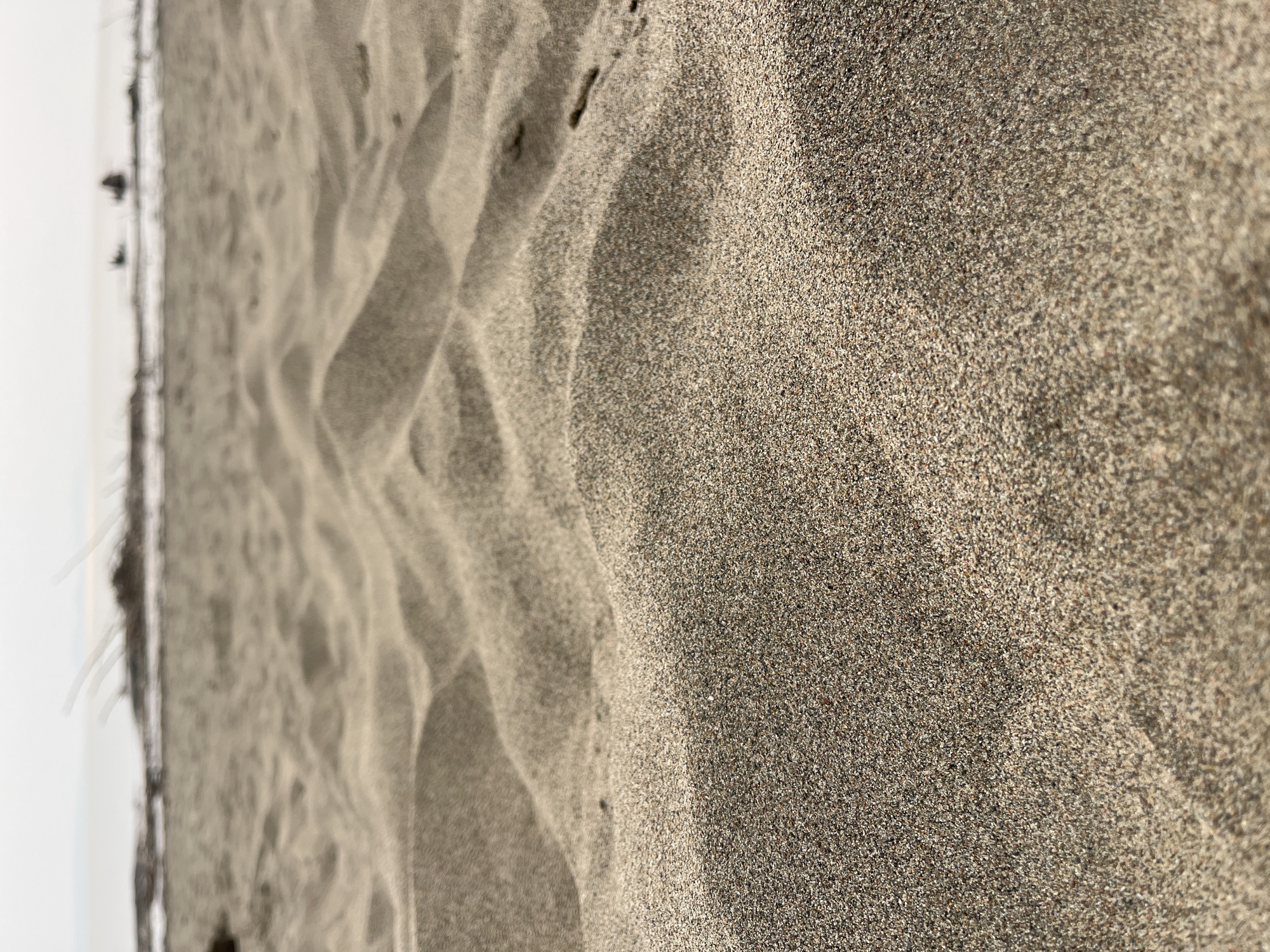 The image shows a close-up, low-angle view of sand dunes on a beach. The sand is light brown and textured, with subtle variations in tone and shadow creating undulating patterns. In the background, a hazy horizon separates the beach from a pale sky, with faint indications of water and some dark, indistinct objects, possibly vegetation or debris, along the shoreline. There are no people or discernible structures in the immediate foreground or midground. The overall impression is one of natural, unoccupied beach landscape. The location is identified as Krasnoturansk, Russia.