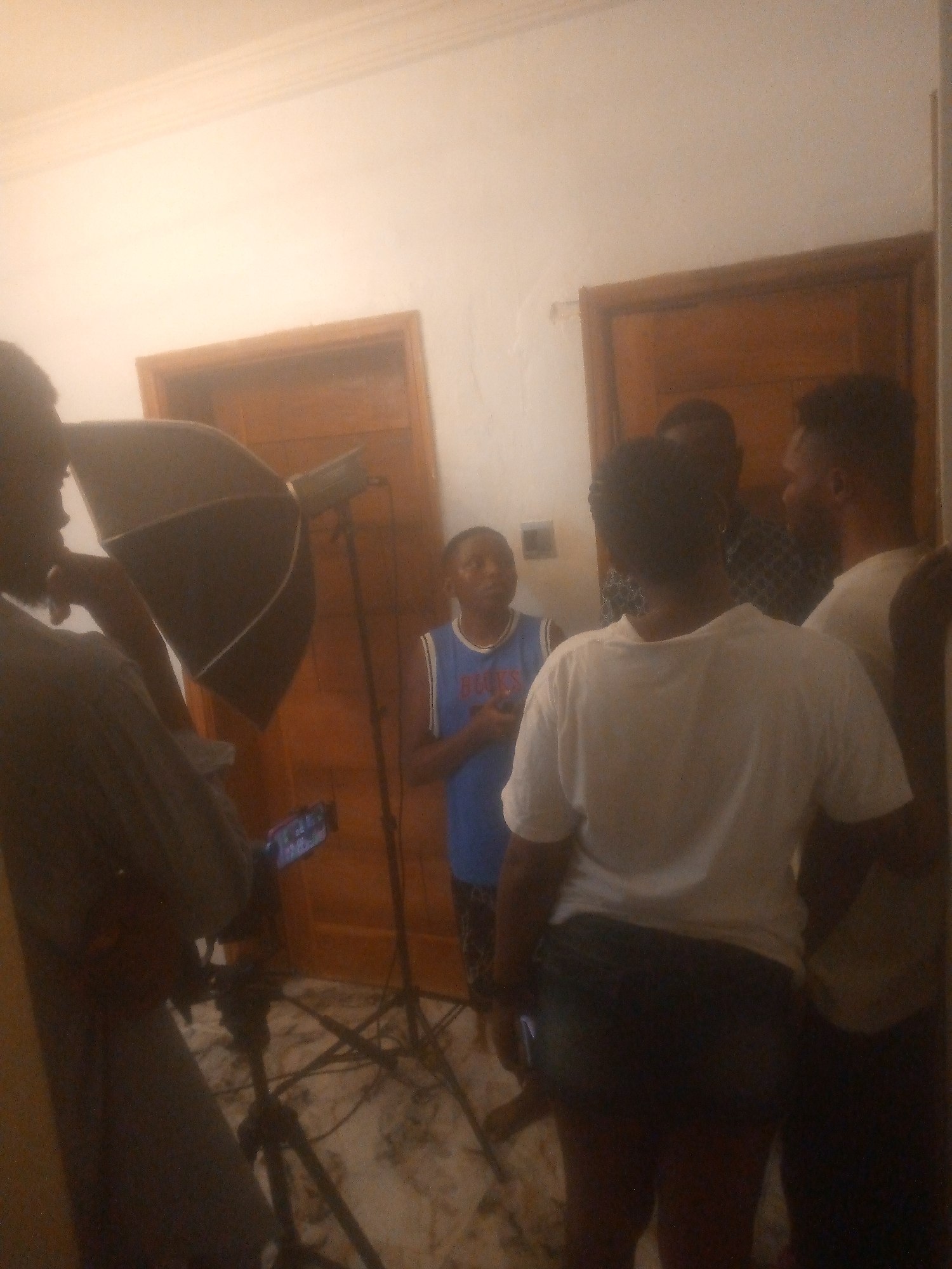 The image depicts an indoor scene, possibly a room in a building in Ikorodu, Nigeria. Several individuals are present, including one person wearing a blue basketball jersey, apparently the subject. The others appear to be associated with a photographic or video shoot. Studio lighting equipment, including an umbrella reflector and a camera on a tripod, are visible. The room has at least two wooden doors. The floor appears to be marble or tile. The overall lighting in the room appears to be a mix of natural and artificial light.