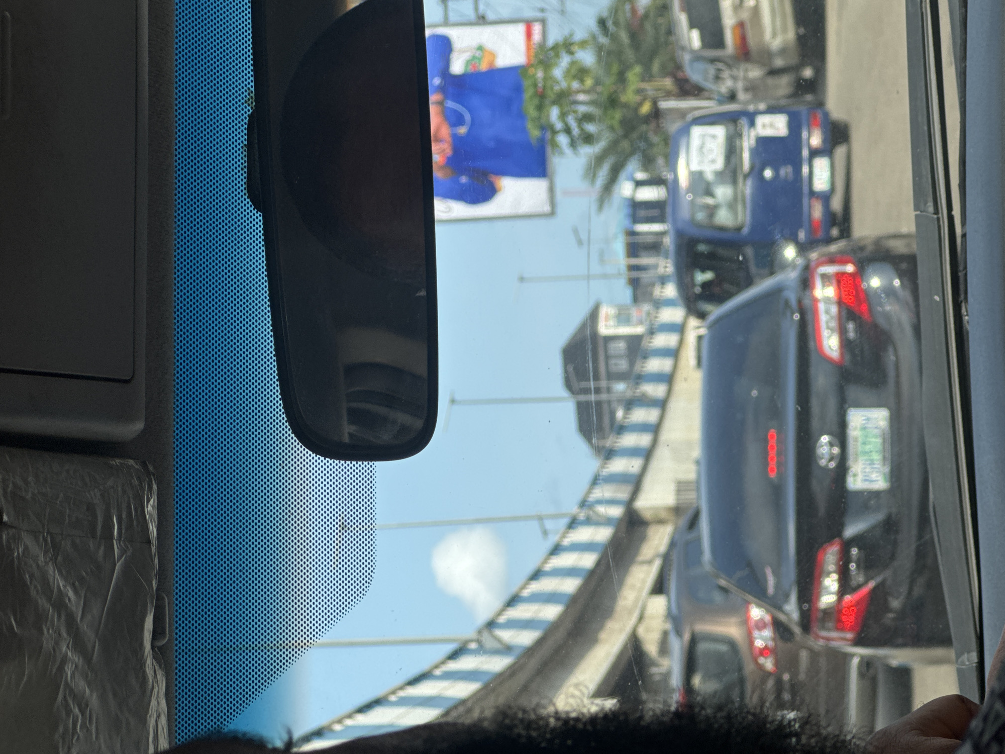 The image, taken from inside a vehicle, shows a traffic jam in Port Harcourt, Nigeria. The view through the windshield is partially obscured by a blue sunshade with a grid pattern and the rearview mirror. In the background, there is an elevated road bridge. Various vehicles including a "keke napep" or auto-rickshaw, are visible in the congested traffic. There is a billboard in the background with an image of a person in blue clothing. The sky is visible through the gaps between vehicles and above the bridge.