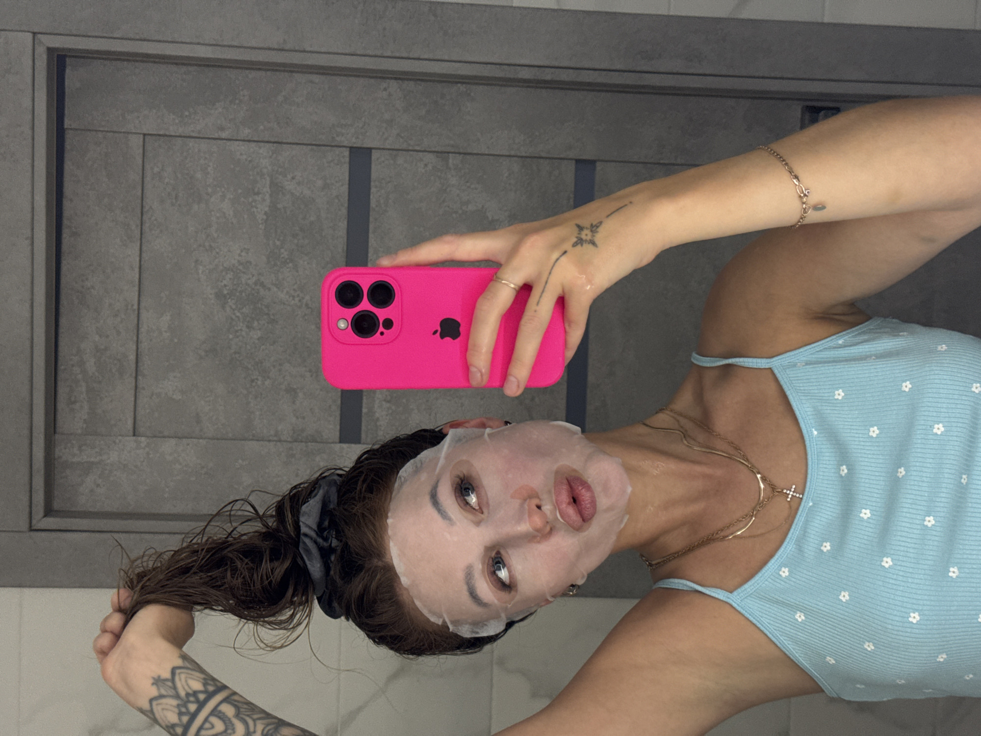 A young woman captures a mirror selfie, seen from an inverted perspective, likely within a modern bathroom setting in Chaikovsky, Russia. Her face is covered by a translucent sheet mask, through which her open eyes and slightly pursed lips are visible. Her dark brown hair, appearing damp or wet, is pulled back and secured with a dark scrunchie. She wears a light blue camisole top adorned with small white floral patterns, and multiple gold necklaces, one featuring a small cross pendant. A delicate gold bracelet is on her right wrist, and a gold ring is on her left hand.
She holds a vibrant hot pink iPhone, identifiable by its triple camera array, flash, and the visible Apple logo on the case. Distinctive tattoos are visible: a small, star-like design on the back of her left hand and an intricate, mandala-style pattern on her right forearm. The background consists of grey, textured panels or tiles, possibly on a wall or cabinet, featuring subtle variations in tone and defined lines. The overall impression is of a personal moment, likely a skincare routine.