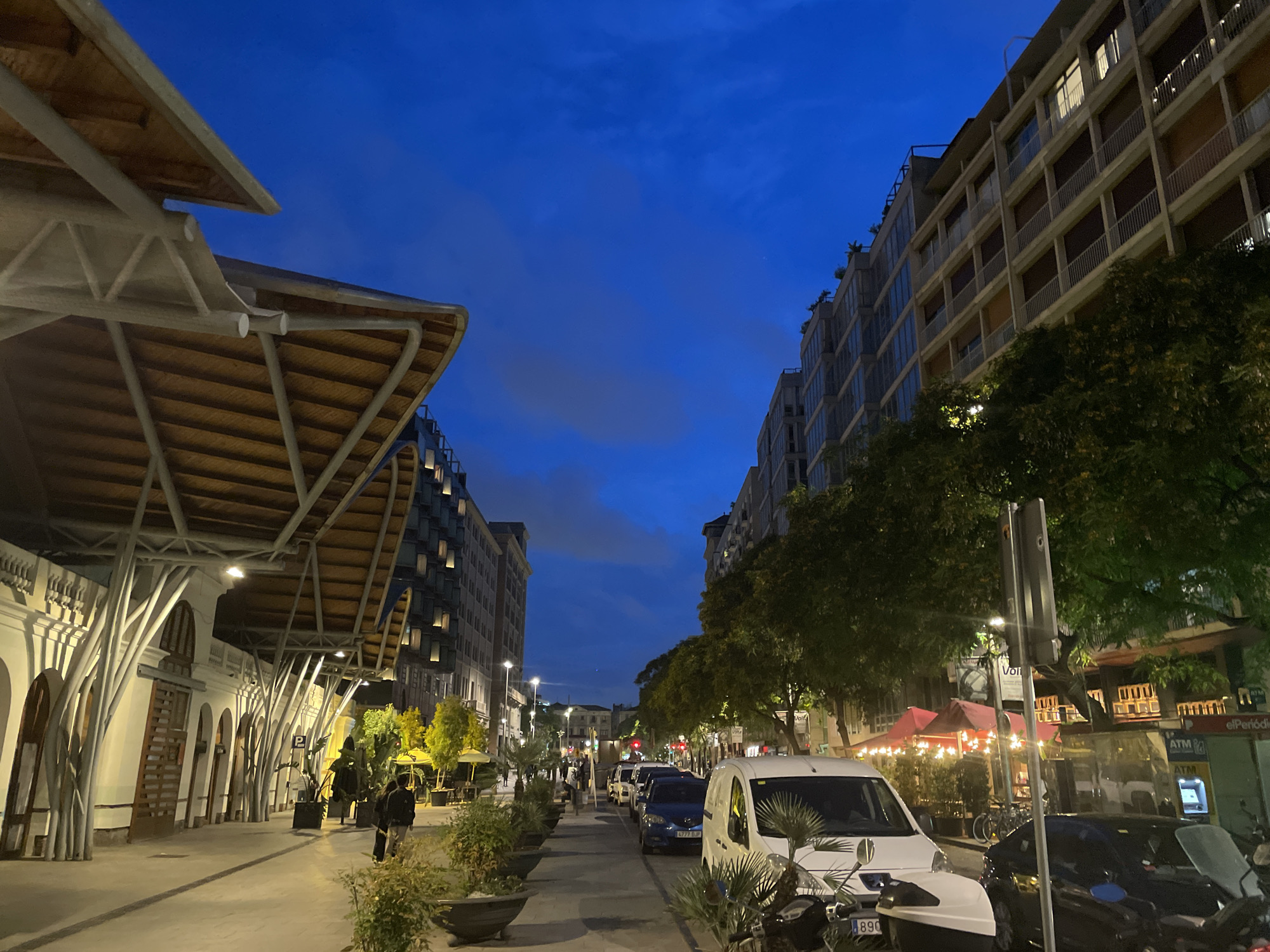 The image depicts a street scene at dusk or early night in Barcelona, Spain, specifically on Carrer del Comte d'Urgell, running alongside the Mercat de Sant Antoni. The sky is a deep indigo blue with faint clouds.
On the left, the prominent Mercat de Sant Antoni features a distinctive modern canopy with an undulating wooden underside, supported by numerous white, tree-like metal pillars. Behind this structure, the traditional market building with light-colored arched entrances and windows is visible. A wide pavement extends along the market, where several individuals are walking. Planters containing green and some yellowing foliage line the edge of this pedestrian area.
To the right, a multi-story modern apartment building with numerous balconies and rectangular windows dominates the background. A row of trees with dense green foliage lines the street, partially obscuring the lower floors and establishments. Below the trees, outdoor dining areas with red awnings and string lights are visible, with people seated. An ATM is located near a visible "elPeriódico" sign.
The street itself is a one-way thoroughfare with vehicles parked along both sides. In the foreground, a white van (with a partially visible Spanish license plate ending in "890") is parked. Further down, a blue car (Spanish license plate "4777 DJF") and other vehicles are present. Streetlights illuminate the pavement and road, complementing the artificial lights emanating from buildings and commercial establishments. A blue "P" parking sign is visible on the left side of the street.