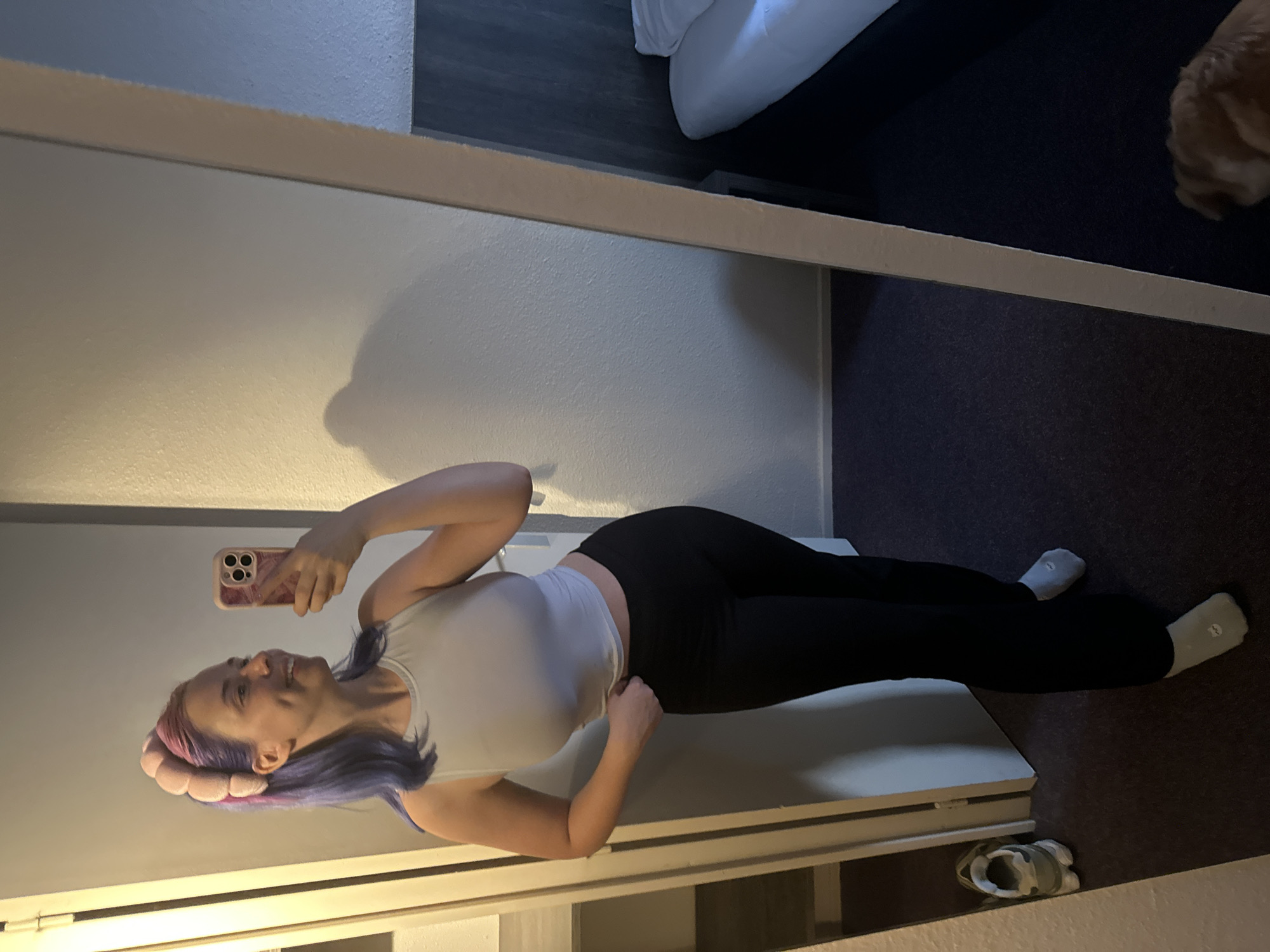 A woman with purple and pink hair is posing in a full-length mirror in a room. She is wearing a white top and black leggings, along with white socks. She is holding a cell phone in her left hand, presumably taking the picture. A bed with white linens and a brown dog are visible in the upper right corner of the mirror's reflection. A pair of shoes is near the bottom right corner of the image. The location is indeterminate and there is no basis in the image to suggest the location is Ramonville-Saint-Agne, France.