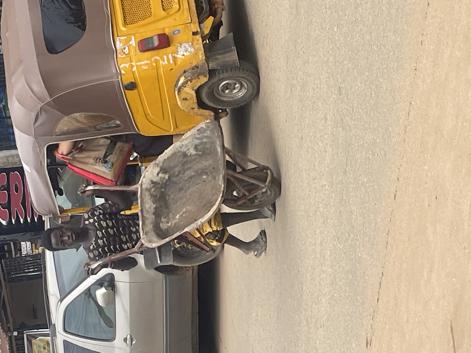 A young man is pushing a wheelbarrow on a paved street in what appears to be an urban area. He is wearing a dark t-shirt with a repeating pattern of small white and black shapes and yellow and black patterned shorts. He has on dark sandals. The wheelbarrow has a metal basin and two handles, and it is being pushed from behind. To the right of the man is a yellow motorized tricycle, commonly used as a taxi in Nigeria. The tricycle has a brown canopy and a visible license plate with white lettering. Behind the man and to the left is the rear of a silver SUV. The background shows building facades, suggesting a commercial or residential area. The overall scene depicts a daily activity, possibly related to transportation or labor, in a Nigerian city like Abuja.