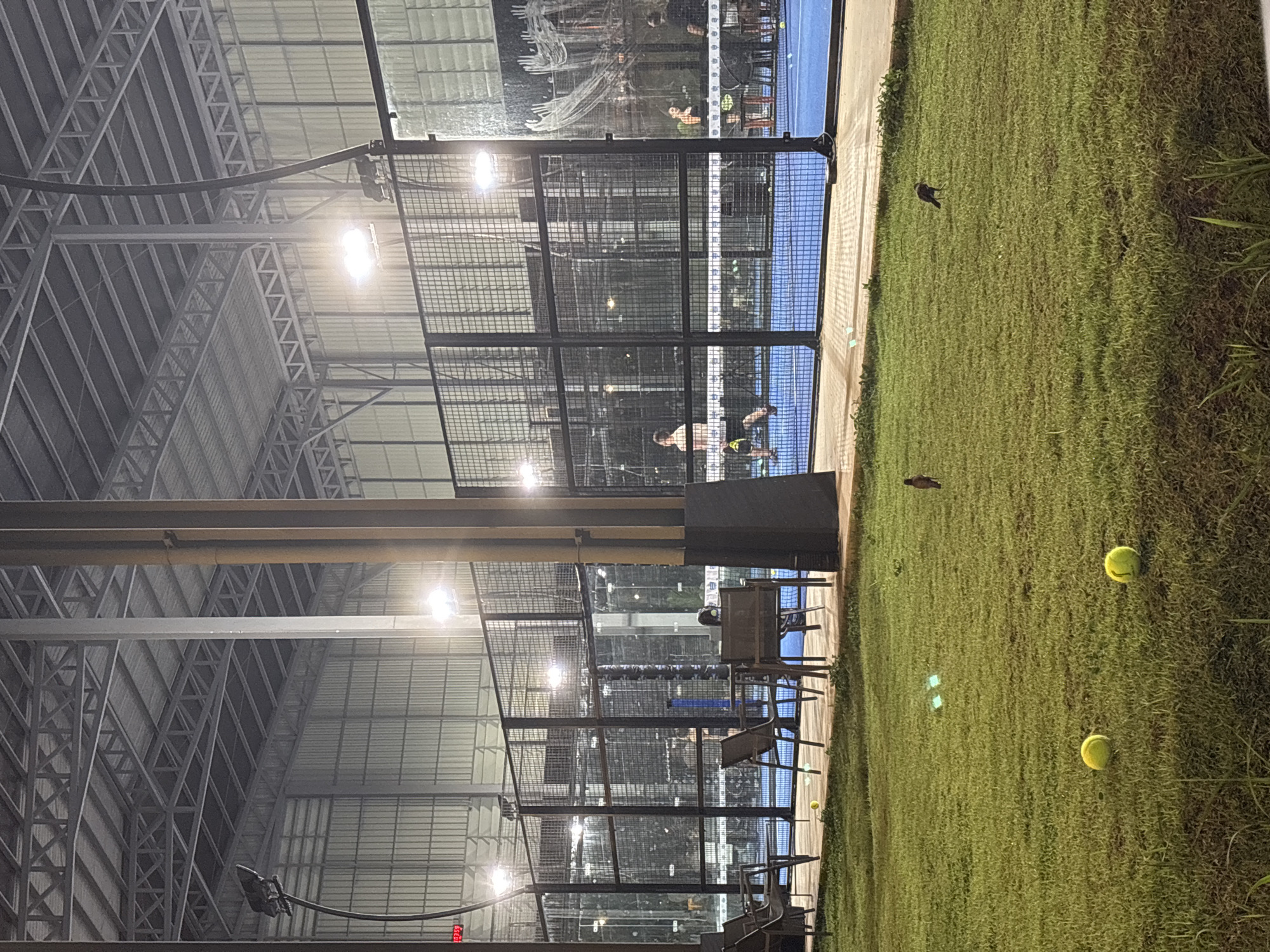 The image depicts an indoor sports complex in Choeng Thale, Thailand, likely for paddle tennis or a similar sport. The space is enclosed by a tall, metallic structure with artificial lighting. A paddle tennis court with glass walls is visible, with people playing on the court. Outside the court, there is a grassy area with tennis balls scattered on the ground. Benches and chairs are placed next to the court for spectators. The scene appears to be taken at night or in the early morning, given the presence of artificial lighting and the dim ambient light.