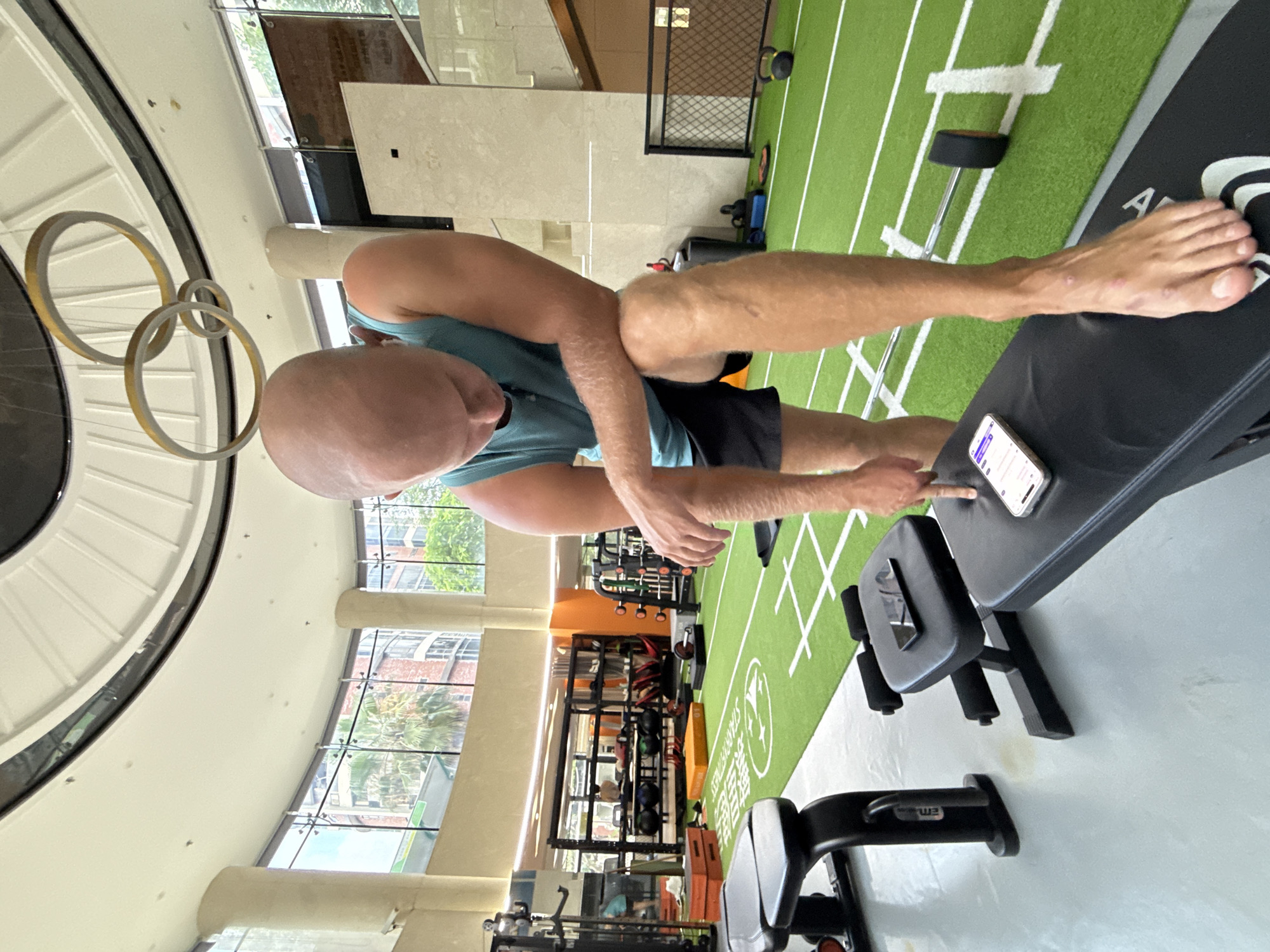 A bald man wearing a teal tank top and black shorts is standing in a gym, leaning forward with his elbow on his knee. He is pointing at a smartphone on a black bench. Another smartphone is on a different black bench in the foreground. The gym has a green turf floor with white lines, weight racks, and other exercise equipment. The background shows large windows with views of buildings and trees. The location is identified as Haizhu District, China.