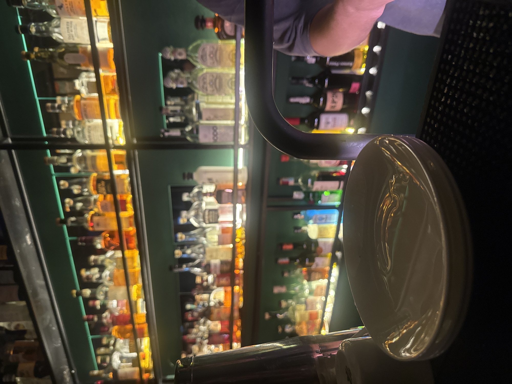 The image depicts a bar interior. A shelf filled with liquor and wine bottles is visible in the background, blurred due to shallow depth of field. A metal bar railing and a stack of white plates are in the foreground. A person wearing a gray shirt is partially visible at the top of the frame. The location is potentially a bar in Budva, Montenegro.