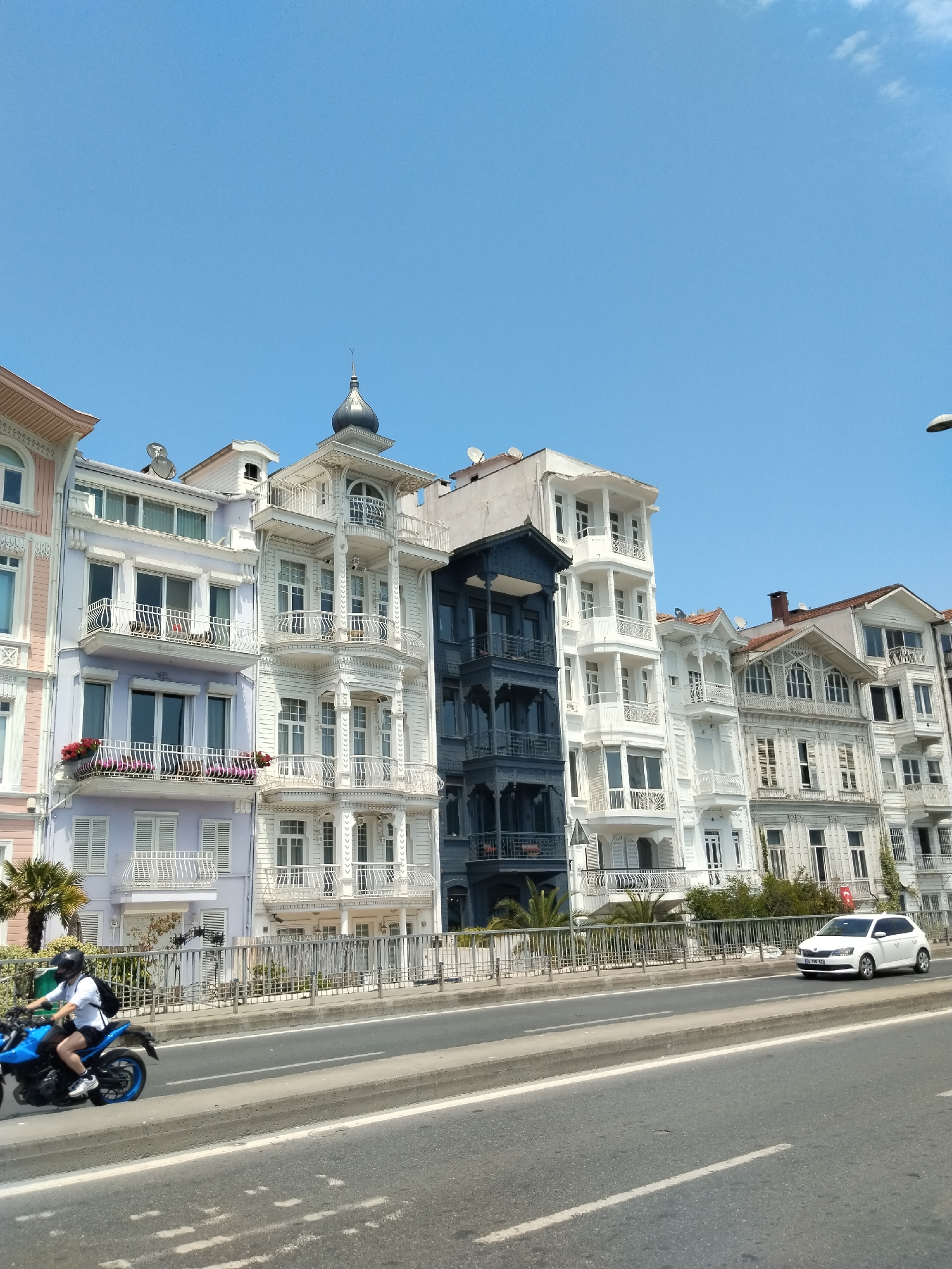 A bright, sunny street scene unfolds under a clear blue sky, showcasing a row of distinct, multi-story buildings lining one side of a multi-lane asphalt road. These ornate structures exhibit a variety of facades, including pale pink, lavender (with balconies adorned with red flowers), and several white buildings featuring intricate wooden details, multiple balconies, and bay windows. A notable dark blue/grey wooden building stands out in the middle of the row. Some rooftops bear satellite dishes, and one white building is topped with a decorative dome-like spire.
A metal barrier fence separates the buildings from the roadway, with glimpses of palm trees and other greenery visible behind it. On the road, a person wearing a black helmet, white long-sleeved shirt, black shorts, and a black backpack rides a blue motorcycle with blue wheels in the foreground, traveling left to right. Further down the road on the right lane, a white Skoda Fabia car is visible, its license plate reading "34 TDR 929". The clear blue sky and bright sunlight indicate a mid-day or early afternoon setting. The architectural style, reminiscent of traditional Bosphorus "yalı" houses, strongly suggests a location in Istanbul, consistent with Beşiktaş, Turkey.