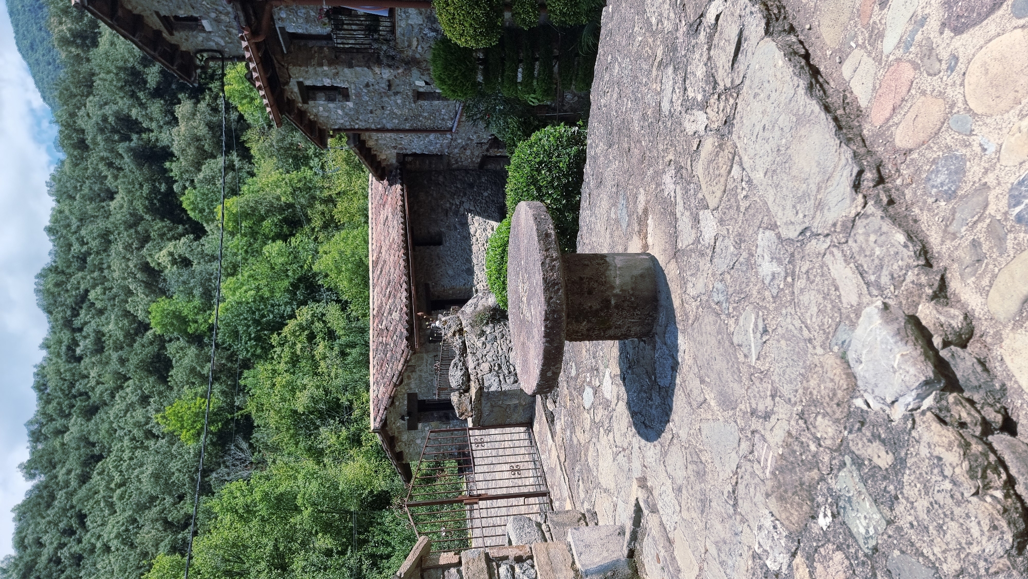 The image depicts an outdoor stone courtyard in front of rustic stone buildings, with a dense, green forested hillside in the background. A circular stone table with a thick pedestal stands in the foreground, casting a shadow on the cobblestone pavement. To the left, a wrought-iron gate is set into a stone wall. The buildings appear to be constructed from rough-hewn stones with tiled roofs. Lush green bushes are visible around the buildings. The scene suggests a rural or historic setting, possibly a courtyard of an old estate or village.