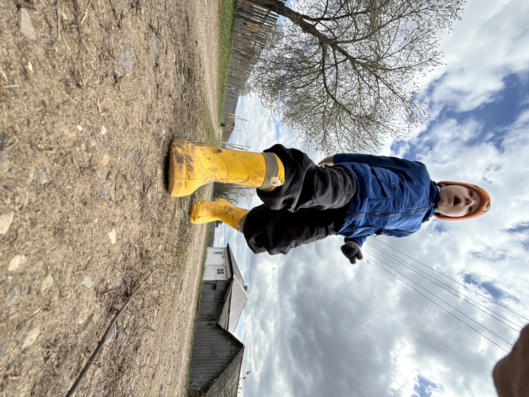 A low-angle shot captures a young child outdoors in Nizhnyaya Chermoda, Russia. The child, possibly a boy, is centered in the frame, looking directly upwards at the camera with his mouth slightly open, conveying an expression of surprise or speaking. He is dressed for cool weather, wearing a vibrant blue zip-up jacket, dark pants, and a bright orange knit hat. His feet are clad in distinctive, muddy yellow rubber boots, indicating recent outdoor activity in wet or dirty conditions.
The setting is a rural or village environment. The ground beneath the child is a dirt and gravel path. In the background, to the left, a long wooden fence or structure stretches into the distance, and further back, traditional wooden houses or buildings with dark roofs are partially visible. A large, bare-branched tree, possibly in early spring before leaf-out, dominates the upper left portion of the background, its branches silhouetted against the sky.
The sky is partly cloudy, with significant patches of white clouds and glimpses of blue, suggesting it is daytime. Several utility lines crisscross the sky on the right side of the image. The overall impression is one of a child enjoying the outdoors in a rustic, possibly remote, location. There is no visible text in the image.