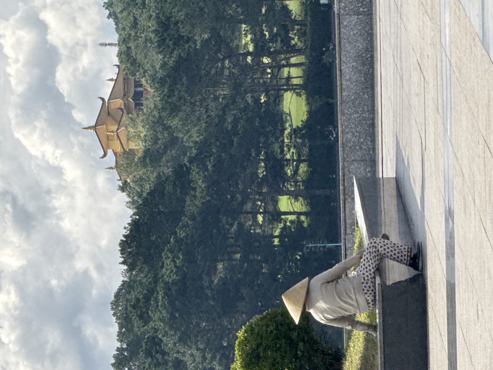 The image depicts a person wearing a conical hat, seated on a concrete bench. The individual is viewed from the back, and is wearing a light-colored jacket and patterned pants. Behind the person, a dense line of dark green trees stretches across the midground. In the background, a golden-roofed building with traditional architecture is visible, perched on a hillside. The sky above is filled with dramatic, cloudy formations. The foreground consists of a paved area. This scene is likely located near a park or a scenic viewpoint in Da Lat, Vietnam, possibly overlooking a forested area and a temple or pagoda.
