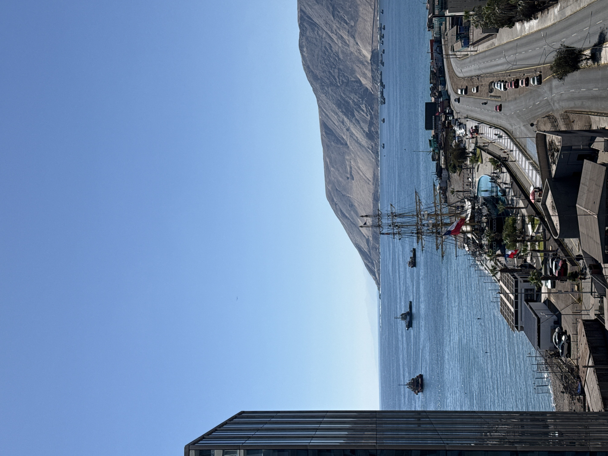 The image depicts a sunny day in Iquique, Chile, showcasing a coastal cityscape with a prominent hill in the background. In the foreground, a modern glass building stands to the left, reflecting the blue sky and surrounding environment. The middle ground features a harbor with several boats, including a large sailing ship docked near a waterfront area. A paved road runs along the coast, with cars and a few pedestrians visible. The ocean stretches to the horizon, meeting the base of the arid, mountainous terrain that dominates the background. The scene suggests a vibrant port city with a mix of contemporary architecture and maritime activity.