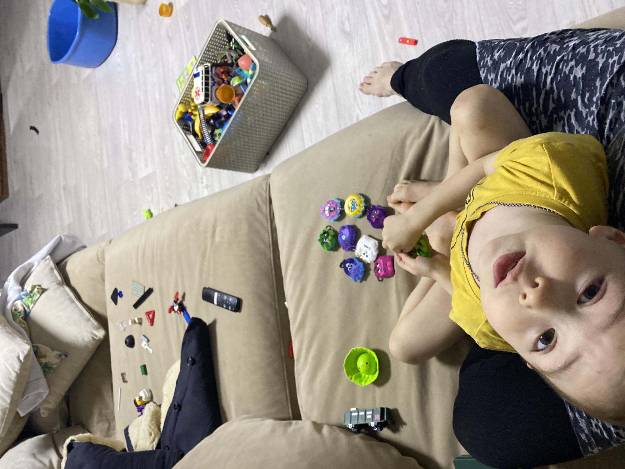 The image depicts an interior scene. A child wearing a yellow shirt is lying on a beige sofa with their face towards the camera. An adult's legs are visible beside the child. The sofa is covered with toys including small, colorful figures and Lego pieces. Other objects on the sofa include a green bowl, a toy train, and a remote control. A gray basket full of toys is on the floor beside the sofa. A blue bucket sits in the background. No information on the location being in Noviy, Russia, can be determined from the image alone.